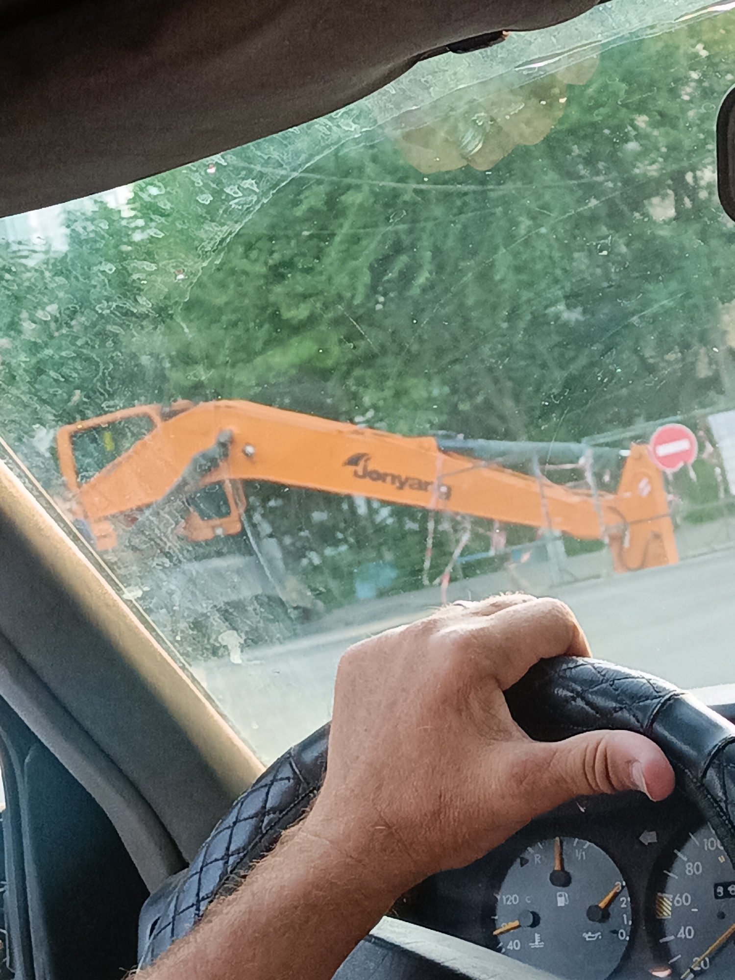 A person's hand grips a steering wheel covered in a black, diamond-patterned material. The hand is tanned and appears to be a male's. The vehicle's dashboard is visible, with a speedometer showing approximately 40 km/h and a fuel gauge indicating low fuel. In the background, seen through a dirty windshield, is an orange construction vehicle, possibly an excavator arm, with the word "Jonyang" visible on it. There are also some traffic signs and trees visible. The overall scene suggests a person is driving a vehicle in an urban or suburban environment.