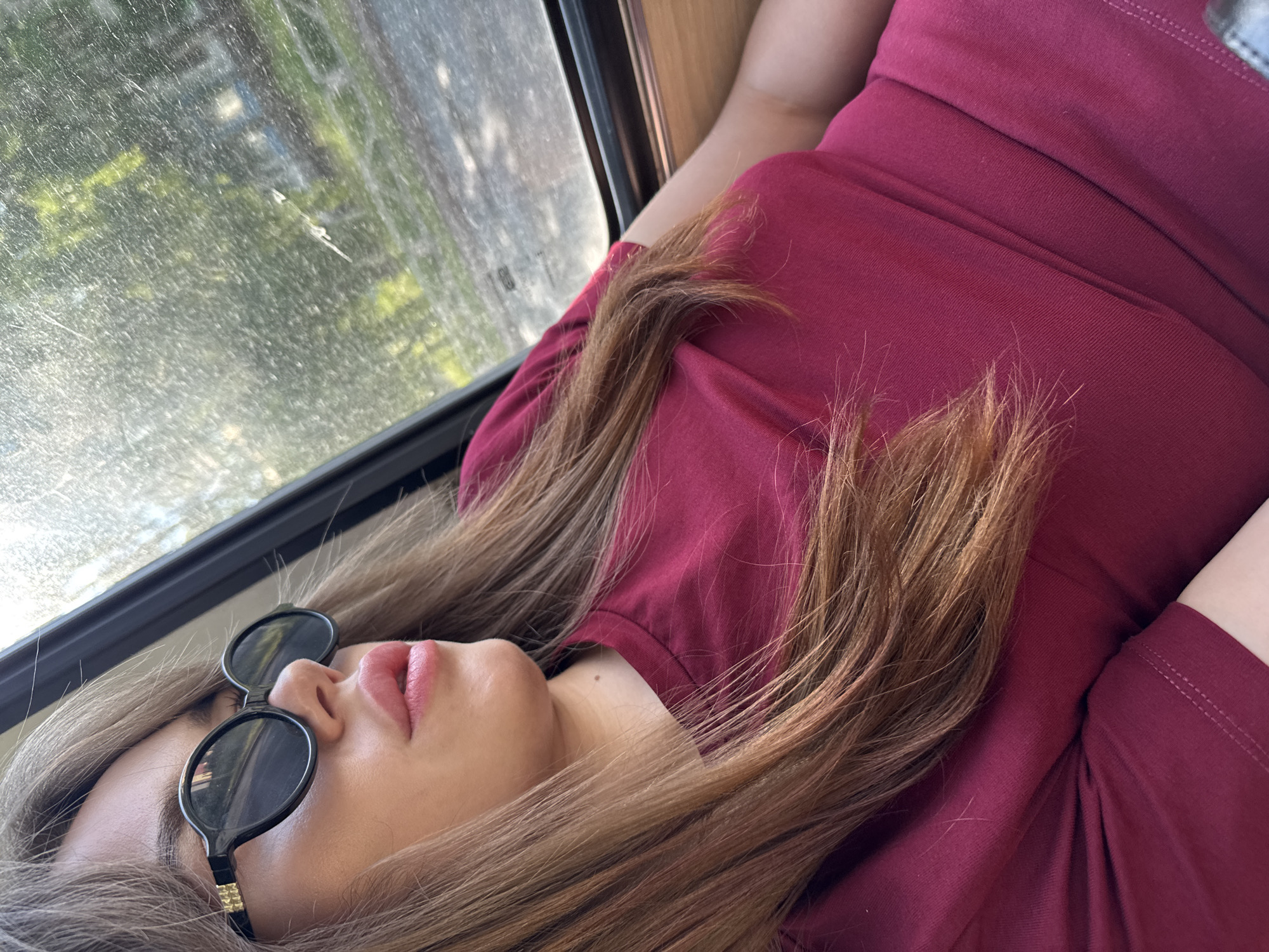 A woman with long, light brown hair and full lips is wearing sunglasses and a maroon t-shirt. She is looking out of a window, which is dusty and shows a blurry background of trees and some buildings. The scene appears to be inside a vehicle, possibly a car, as suggested by the window frame and the angle of the shot. The woman is positioned on the right side of the frame, with her head tilted slightly to the left. The lighting suggests it is daytime. The image was taken in the city of Chaikovsky, Russia.