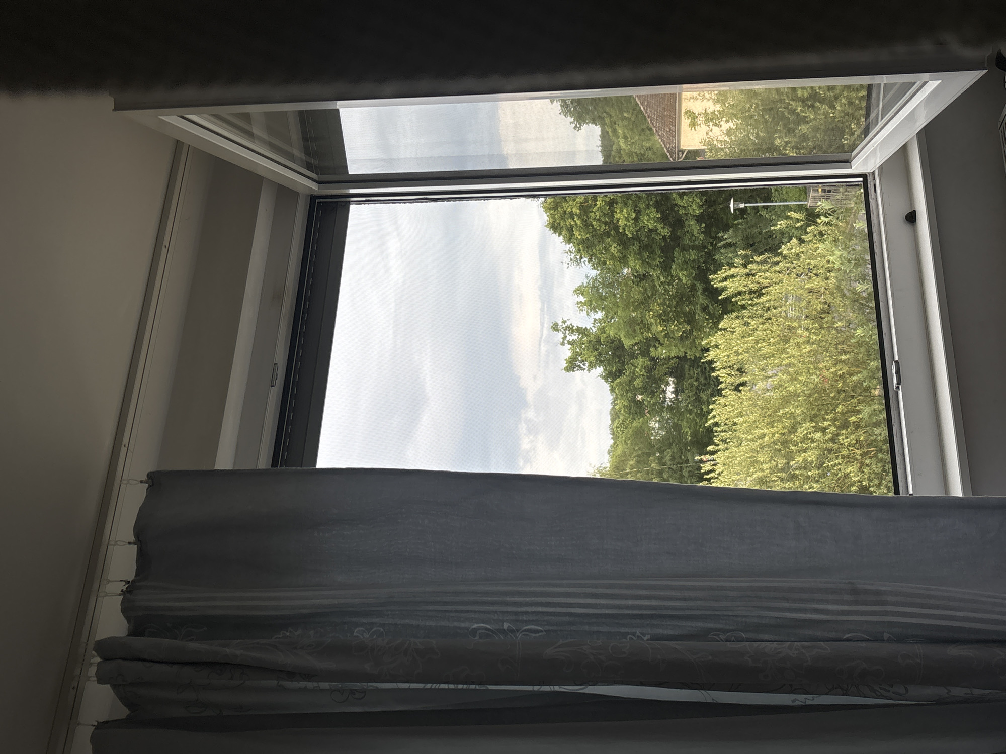 The image shows an interior view through an open window, looking out onto a green, treed landscape with a cloudy sky. A gray curtain is pulled to the left side of the window. The window frame is white, and the outer pane is partially open. Beyond the window, there are dense green trees and bushes. In the background, a building with a tiled roof is visible. The setting appears to be residential. Based on the available information, this view is consistent with a location in Göttingen, Germany, likely a residential area with mature trees. There are no people visible in the image.