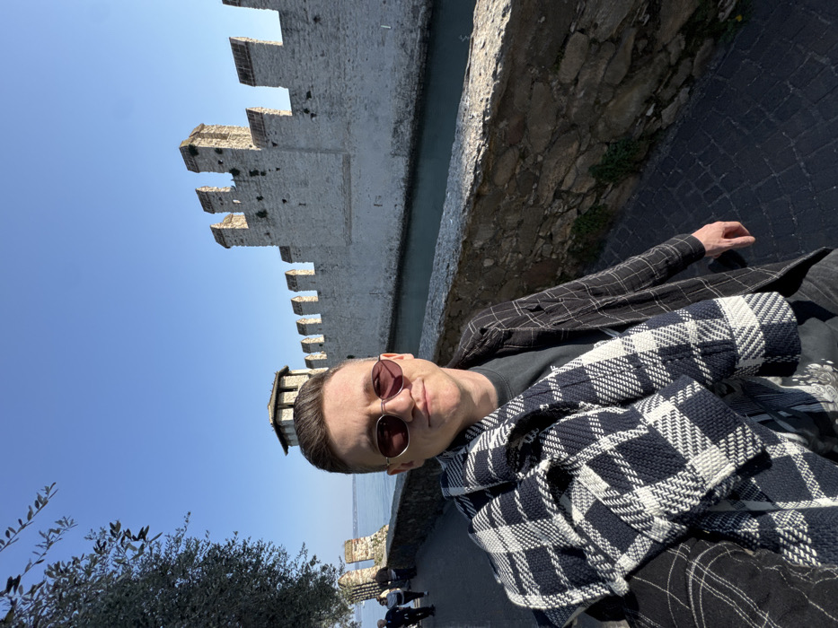 An adult male subject is depicted in a selfie, positioned on a paved walkway alongside the Scaliger Castle in Sirmione, Italy. The subject, a light-skinned male with short brown hair and round, rose-tinted sunglasses, looks directly at the camera with a slight smile. He wears a dark t-shirt under a dark, possibly plaid, long-sleeved shirt or jacket, and a large blue and white checkered/plaid blanket or thick scarf is draped over his left shoulder and arm. To the subject's right and extending into the background is a light grey stone wall of the Scaliger Castle, featuring Ghibelline battlements and a visible tower section with a conical roof on the far left of the wall. A narrow, dark blue-green moat runs directly in front of the castle wall. To the subject's left, a lower stone wall borders a path where several indistinct individuals are observed walking away from the camera. Beyond this path, a large body of water, identified as Lake Garda, is visible under a clear blue sky. Sparse olive tree branches with leaves are partially visible in the upper left foreground. The scene is illuminated by strong sunlight, casting shadows and highlighting the medieval architecture and natural environment.