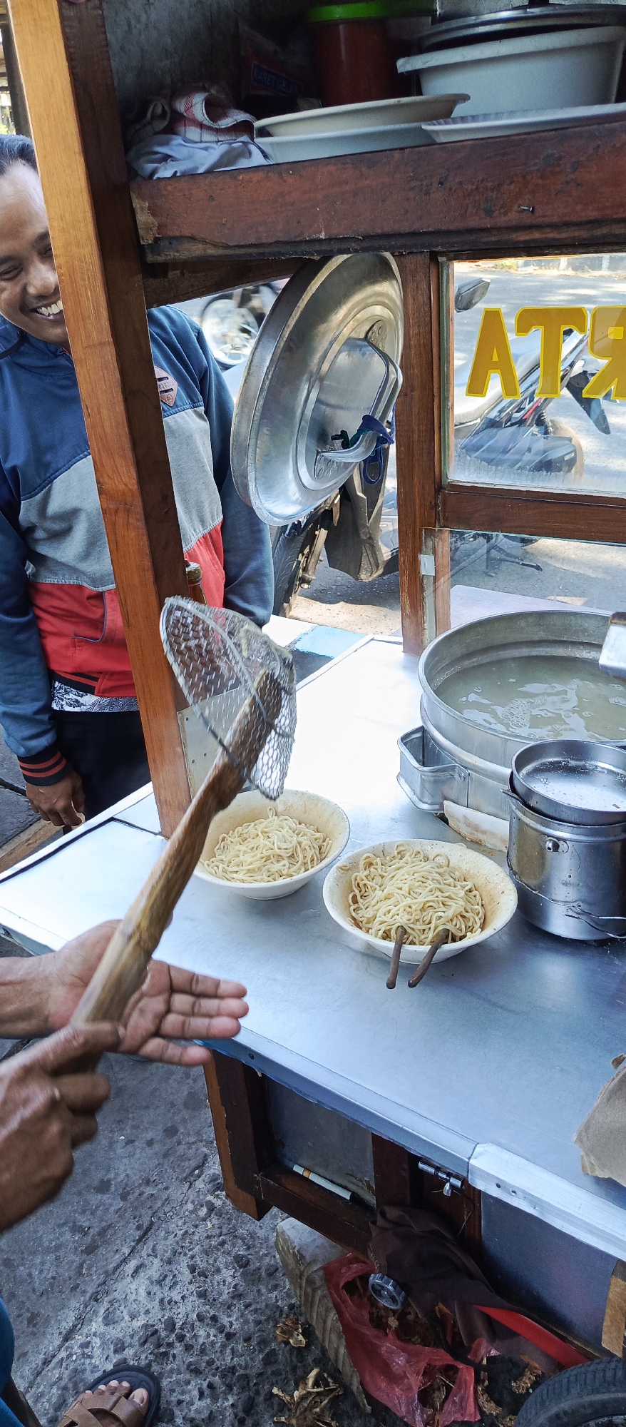 A man smiles as he prepares a meal at a food cart on a paved surface in Wadungasri, Indonesia. The cart is made of wood and metal, with a metal countertop displaying two bowls of noodles and a large pot of broth. The man is using a metal strainer with a wooden handle to serve noodles into one of the bowls. He is wearing a blue, grey, and red jacket. In the background, a motorcycle is visible, and a yellow sign with the letters "AT" is attached to the cart's window. The ground is concrete, with fallen leaves scattered around.