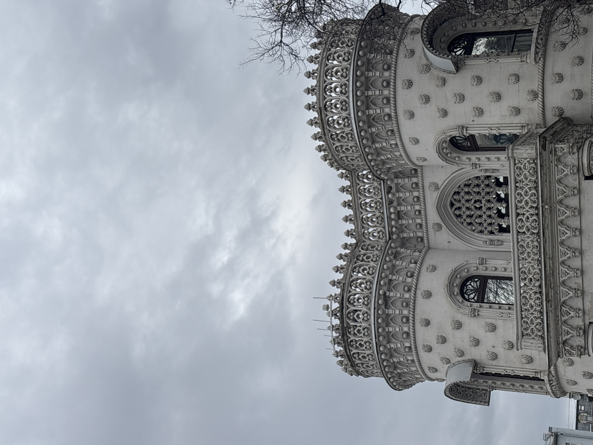 The image shows the exterior of the House of Igumnov, a notable building in Moscow, Russia. The building is decorated with intricate white stone carvings, including shell motifs, gothic-style tracery, and ornate cornices. The sky is overcast. Bare tree branches are visible at the top of the frame.