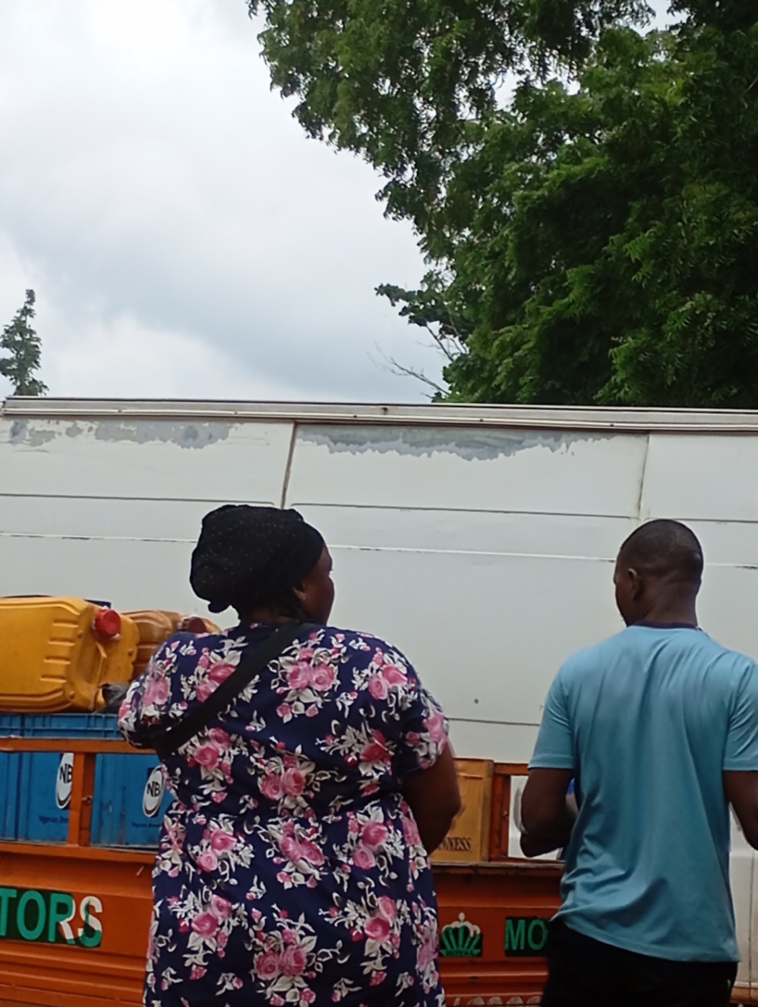 In Shendam, Nigeria, two individuals stand facing a white truck with a partially visible logo on its side. The person on the left, a woman wearing a dark headscarf and a floral patterned dress, has her back to the camera. To her left are several yellow containers. To the right, a man in a light blue t-shirt also faces away from the camera. The truck is parked outdoors, with lush green trees visible in the background against an overcast sky. The lower part of the truck bed is orange and features green lettering that reads "TORS" and "MO".