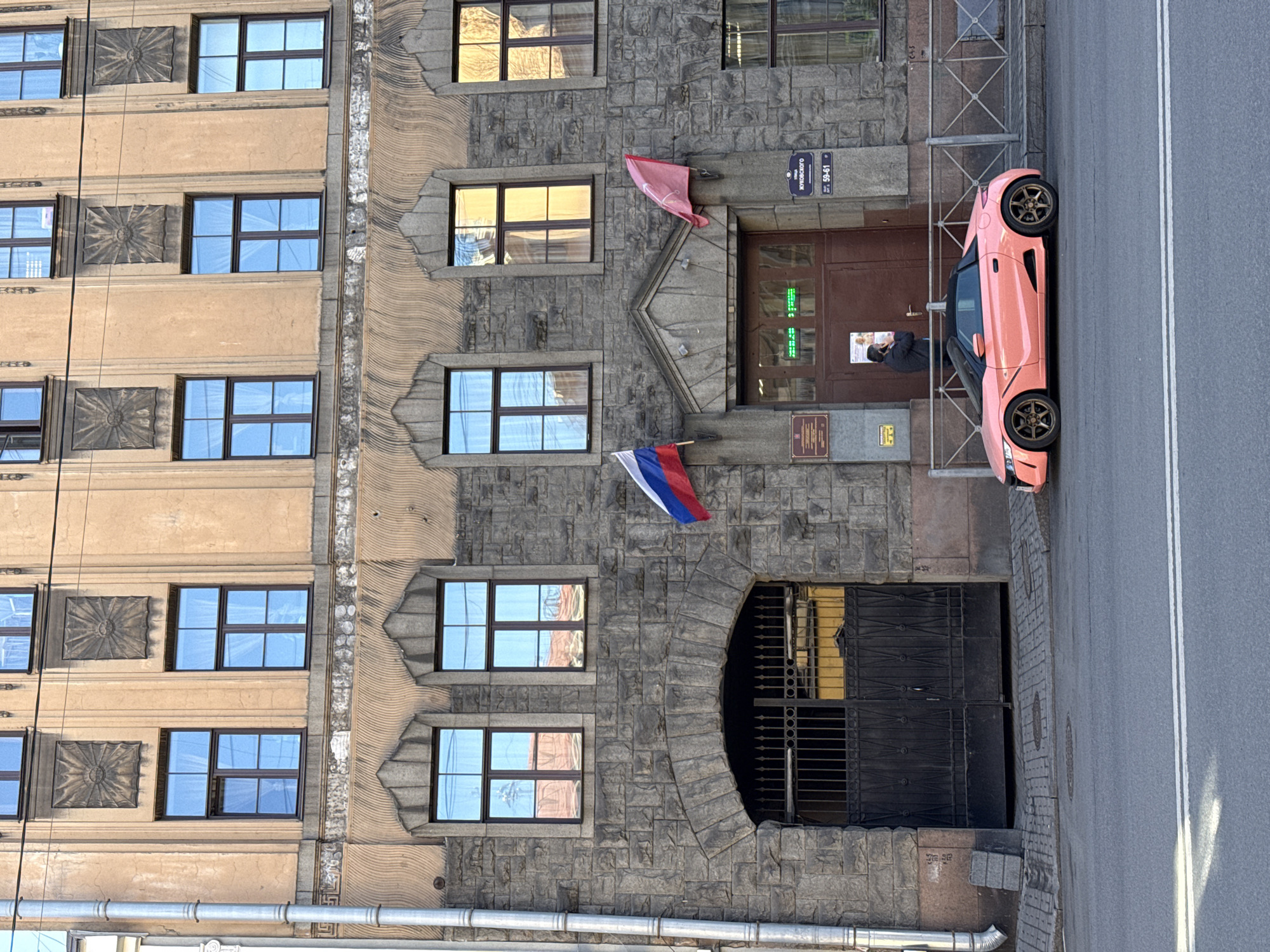 The image shows a building facade in Saint Petersburg, Russia, with a pink car parked on the adjacent street. The building has a mix of architectural styles, featuring stone and stucco sections, with rows of windows and decorative elements. The car, a small two-seater, is parked very close to the building's entrance, which has a sign indicating the address as " Жуковского 59-61". A Russian flag is mounted on the building. A man is visible at the entrance. The street is paved and has white lane markings.