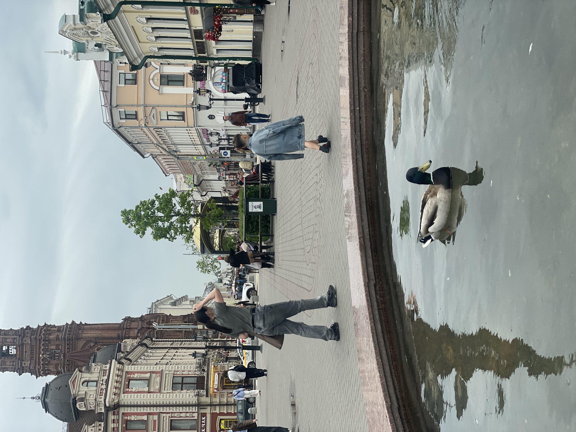 The image shows a street scene in Kazan, Russia. A man wearing a green shirt and gray pants is standing on a brick-paved walkway, holding his head and looking up at a building. He has a bag slung over his shoulder. In the background, there is a building with a brick tower and a dome. The street is lined with buildings, some with ornate facades. There are other people walking along the street. In the foreground, there is a pool of water with a duck swimming in it. The reflection of the buildings can be seen in the water.