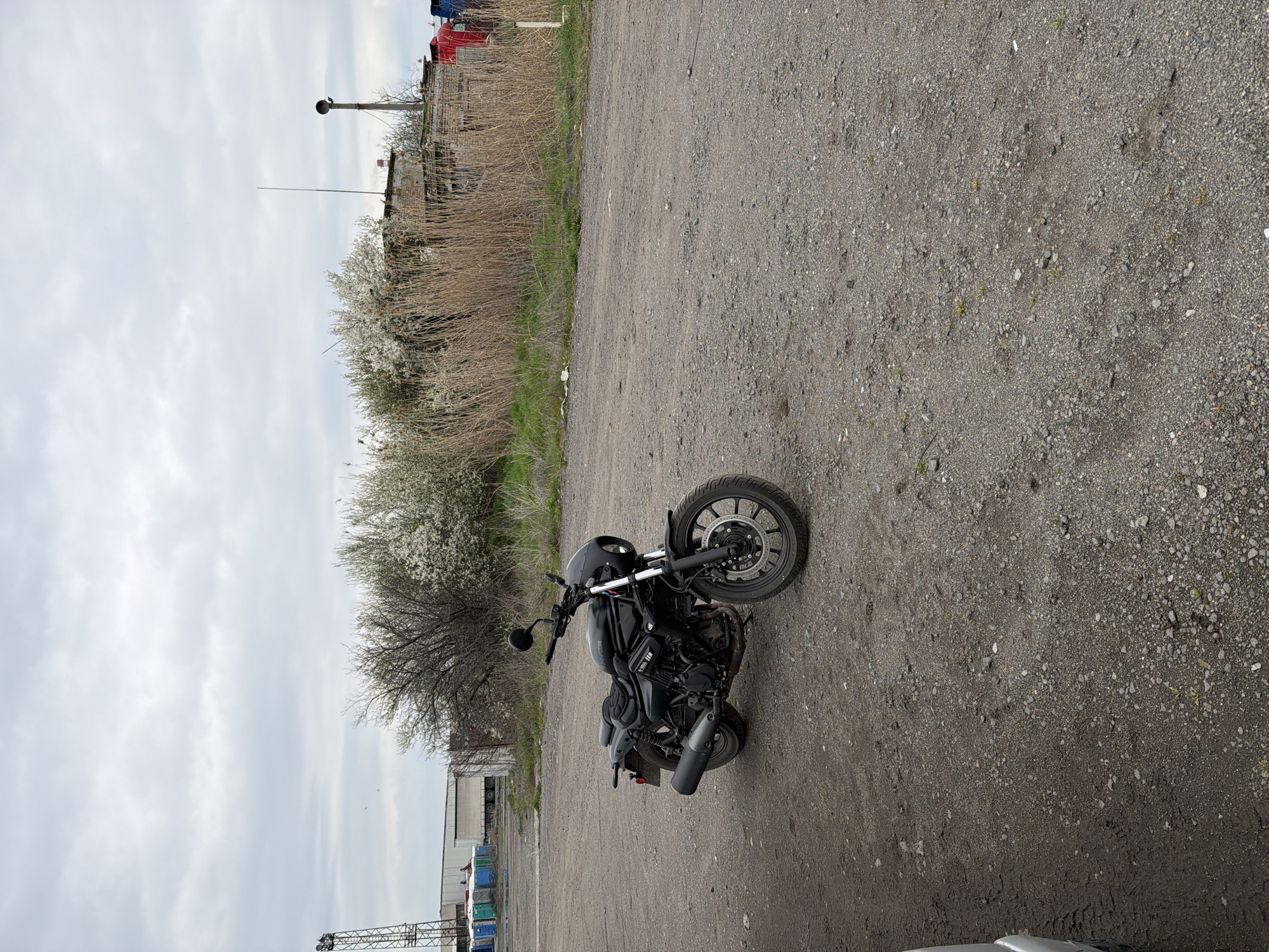 The image shows a matte black motorcycle parked on a gravel lot. The motorcycle is facing right. Behind the motorcycle is a dirt road and vegetation, with buildings visible further in the background. The sky is overcast. The location is likely in Burgas, Bulgaria.
