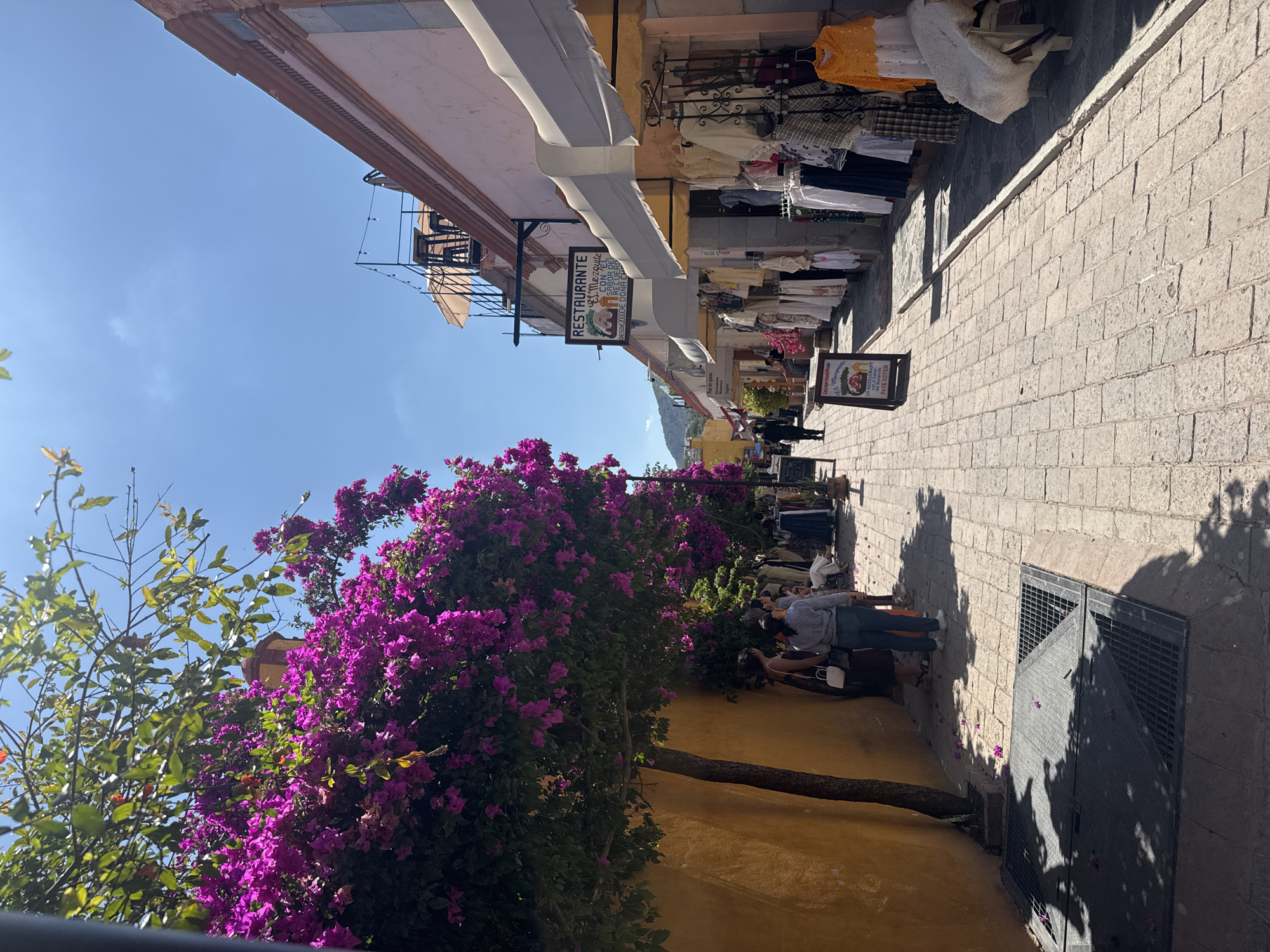 The image captures a street scene in Bernal, Mexico. The camera is positioned looking down the street, with the buildings on either side converging into the distance. On the left side of the street, there's a wall covered in purple bougainvillea flowers. A small tree trunk extends along the yellow-painted wall. A few people are on the street, one leaning against the wall and another appears to be taking a picture. Clothing items hang outside shops on the right side of the street, above a cobblestone walkway. A sign indicates a restaurant, "El Mezquite". The sky above is clear blue. A metal grate or door is seen on the right.