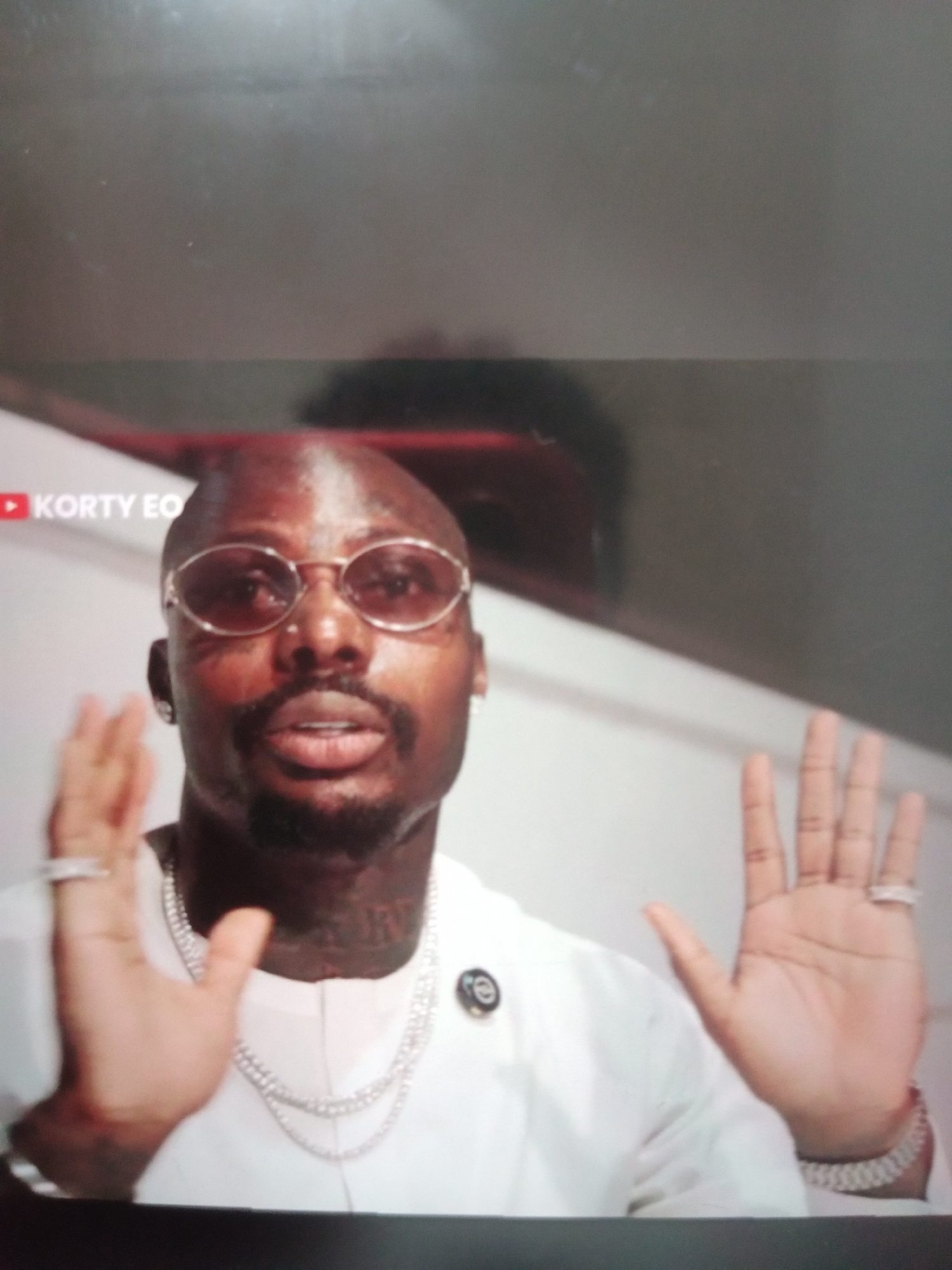 The media file is a close-up shot of a man with a shaved head, wearing round, tinted glasses. He has a goatee and mustache, and appears to have tattoos on his neck. He is wearing a white garment and several pieces of jewelry, including a thick chain necklace, rings on his fingers, and a bracelet. His hands are raised and open, palms facing forward.
The man seems to be speaking or presenting, with his mouth slightly open. The background is blurred, but appears to be an indoor setting with a white wall and a dark ceiling or upper area. There is a subtle red-tinted reflection visible in the upper part of the frame. A logo with the text "KORTY EO" is visible in the upper left corner of the image, accompanied by a red play button icon, suggesting it might be from a video platform.
The lighting suggests it could be daytime, but the indoor setting makes it difficult to determine the exact time of day or weather conditions. The man's expression is neutral, but the gesture of his raised hands could indicate emphasis or a plea. There are no immediate environmental cues to suggest a specific location beyond it being an indoor filming or presentation space. The visible text "KORTY EO" is the only explicit textual information present.