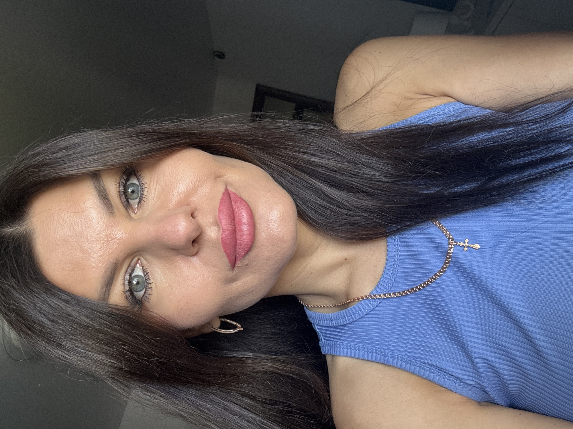 The image is a close-up, waist-up portrait of a woman with long, dark brown hair and light blue eyes. She is wearing a blue ribbed tank top and a gold chain necklace with a cross pendant. She is also wearing gold hoop earrings. Her lips are painted with a matte pink lipstick. The background is dark and out of focus, with a hint of a doorway or mirror visible on the right. The lighting suggests an indoor setting, possibly near a window. There is no information available to determine if the image was taken in Chaikovsky, Russia.