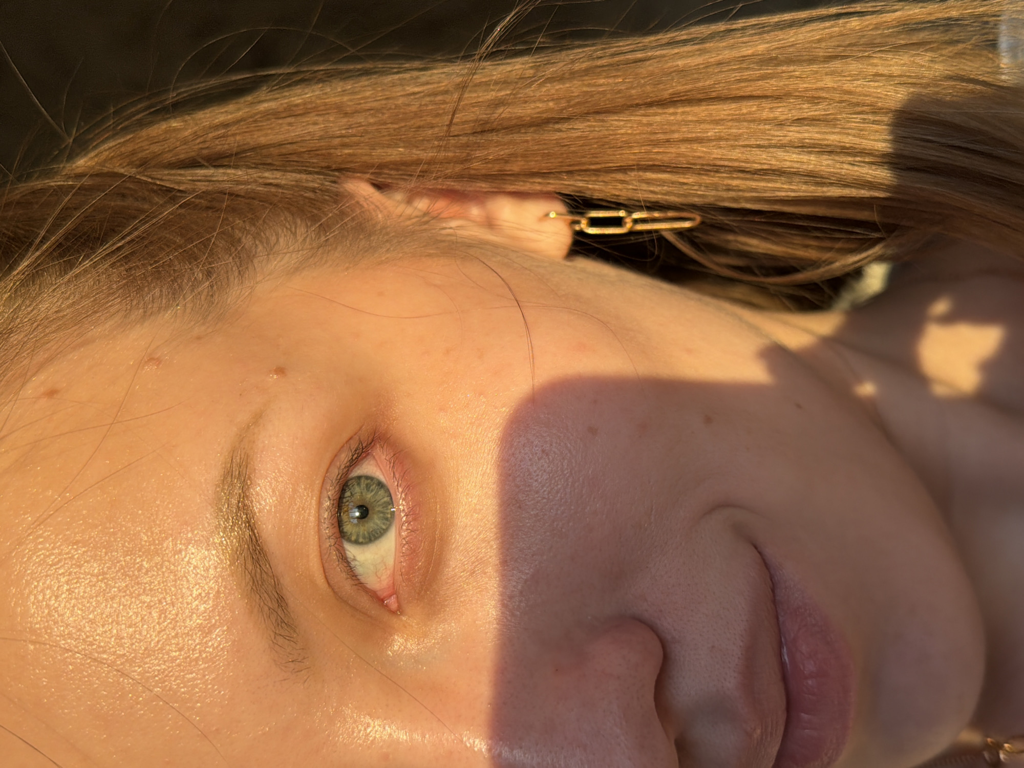 A close-up, tilted view captures the face of a fair-skinned person with green eyes. The light illuminates the skin, revealing fine details such as freckles and pores. The person has light brown hair, with a gold-colored chain-link hair clip visible near the ear. The lower portion of the face, including the nose and lips, is visible. The background is blurry and indistinct. No evidence indicates a location within Reshetnikovo, Russia.