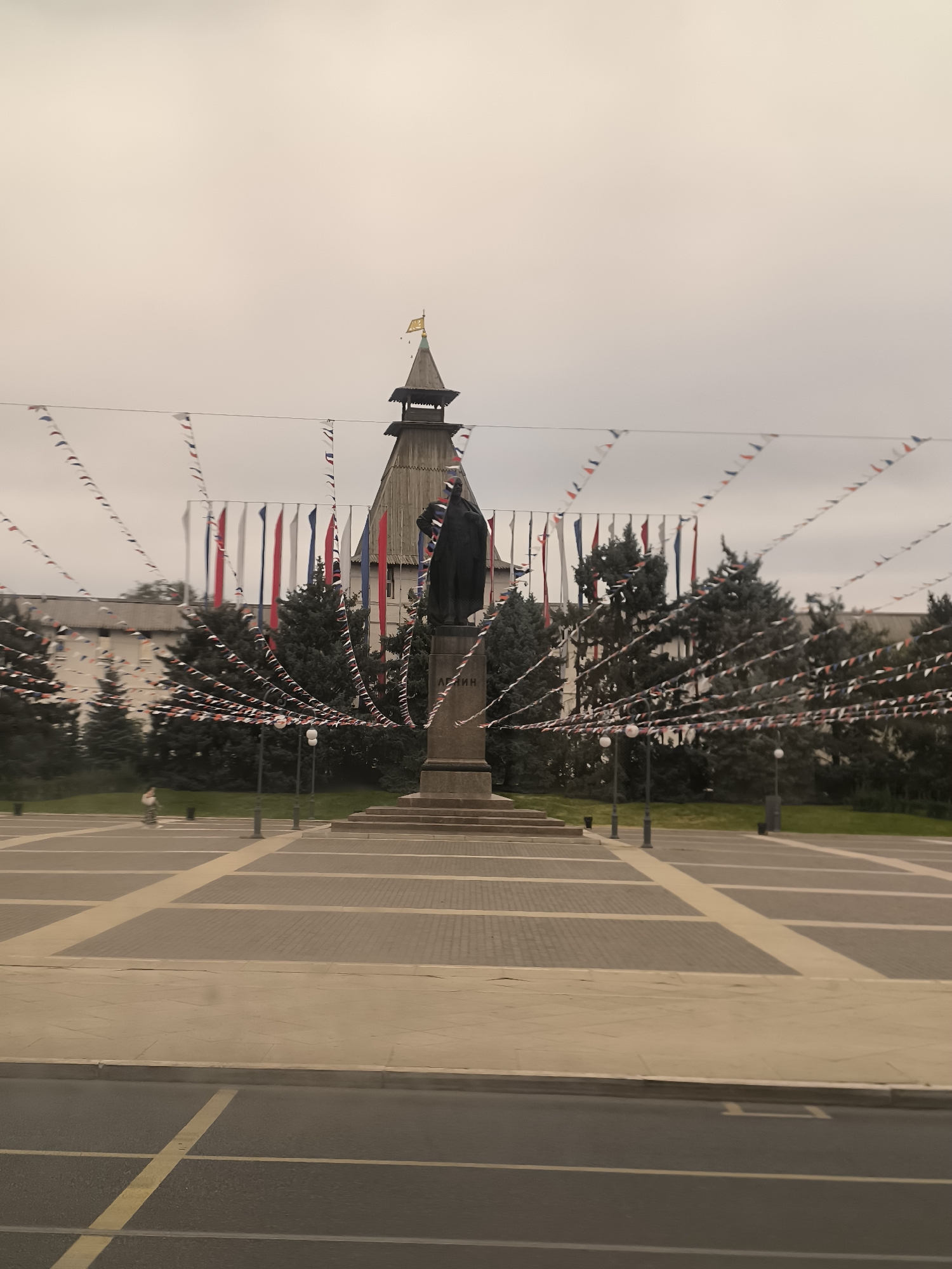 The image shows a wide plaza in front of a historical building in Astrakhan, Russia. A statue of a man in a long coat stands on a pedestal in the center of the plaza, with the inscription "LENIN" visible on the base. The plaza is paved with square tiles in varying shades of gray and beige, forming a geometric pattern. A street with yellow markings is in the foreground. Rows of trees line the background, behind the plaza. The sky is overcast. Numerous strings of small triangular flags in red, white, and blue are strung across the plaza, creating a festive atmosphere. Behind the statue, a multi-tiered wooden tower with a pointed roof and a golden weather vane is visible, attached to a large, light-colored building. The overall scene depicts a public square in front of a significant landmark, decorated for an event.