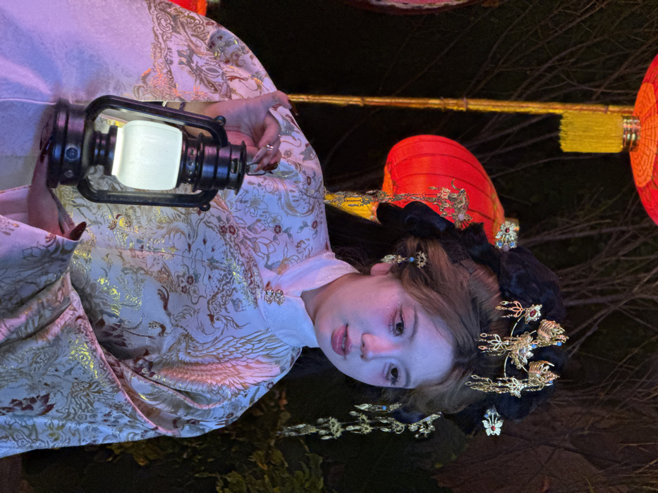 An East Asian female is depicted outdoors at night in Yuzhong District, China, holding an illuminated black lantern. She wears an elaborate traditional Chinese costume, specifically an off-white garment with intricate gold and silver patterns featuring phoenix and floral motifs. The garment has a high collar secured by two decorative clasps. Her dark hair is styled in an ornate updo, heavily adorned with gold-colored hairpins, some featuring red and blue accents and delicate dangling chains. Her face is made up with prominent red eyeshadow and glossy, light pink lips. Her right hand, with dark-painted fingernails, holds the black lantern, which emits a soft white glow from its frosted glass globe. The background is dark, showing indistinct tree branches and several large, red traditional Chinese lanterns, one prominently featuring a yellow tassel, suggesting a festive or cultural outdoor setting.