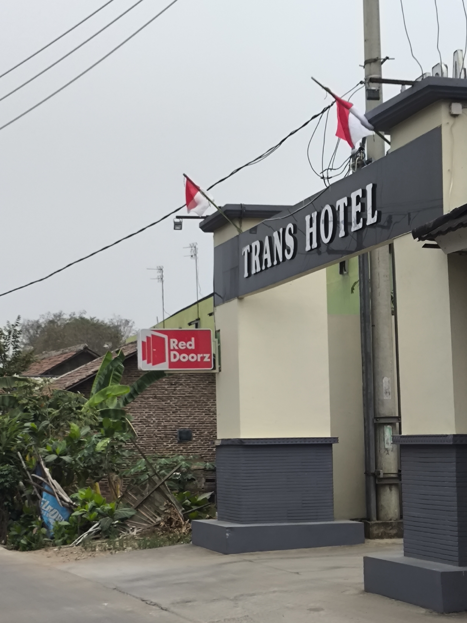 The image shows the entrance to the Trans Hotel in Cilegon, Indonesia. Two Indonesian flags are displayed on top of the hotel entrance, which is a modern design with beige walls and dark grey decorative elements. A sign for "Red Doorz" is visible on the left side of the entrance, indicating a budget hotel chain. Surrounding the hotel is a somewhat unkempt environment with lush greenery, including banana trees, and a brick wall. Electrical wires are visible overhead, and a utility pole stands near the hotel entrance. The scene is captured from a low angle, looking up towards the hotel.