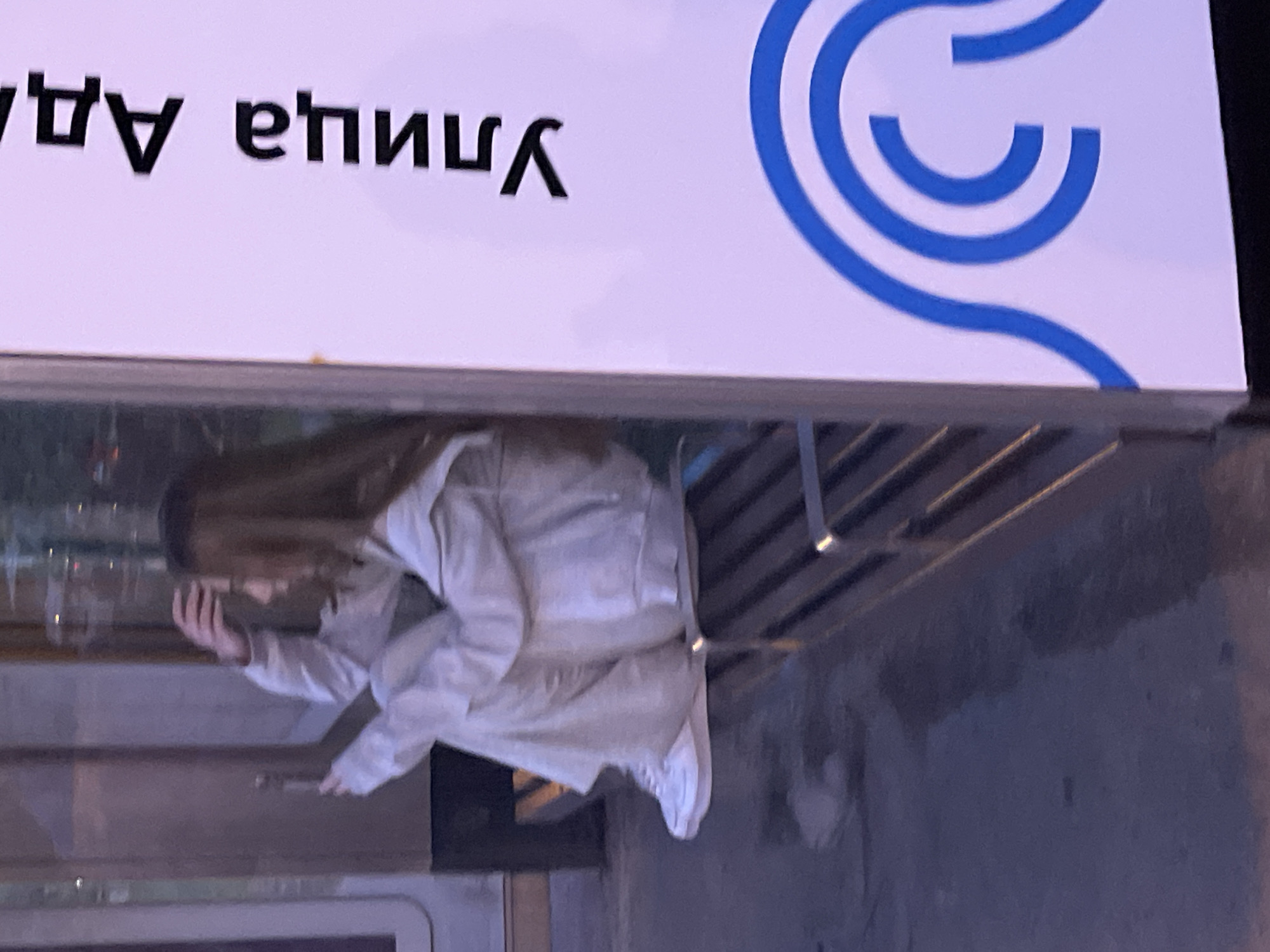The image shows a young woman sitting on a bus stop bench. She is wearing a light-colored tracksuit and white sneakers, and her hand is in her hair. The bus stop has a white panel with blue wavy lines and text in Russian that reads "Улица Адмирала". The scene appears to be a street in Moscow, Russia, possibly in the evening given the lighting. The bench is made of metal slats, and the ground in front of it is wet and reflective.