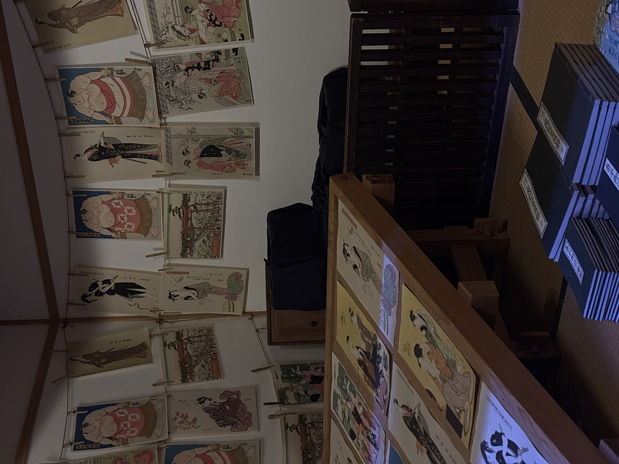 The image depicts an indoor scene within the Sumida Hokusai Museum in Sumida, Japan, showcasing a collection of traditional Japanese ukiyo-e prints. The prints, featuring diverse subjects such as sumo wrestlers, geishas, and landscapes, are displayed on a wall, hung with clothespins on a string, and also laid out on angled wooden display stands. Several stacks of books with Japanese characters on their covers are visible on the floor in the foreground. The environment is dimly lit, with warm ambient light highlighting the artworks and the wooden furniture. A dark fabric is draped over a piece of furniture in the background, partially obscuring it. The overall impression is one of an art exhibition, with an emphasis on historical Japanese art.