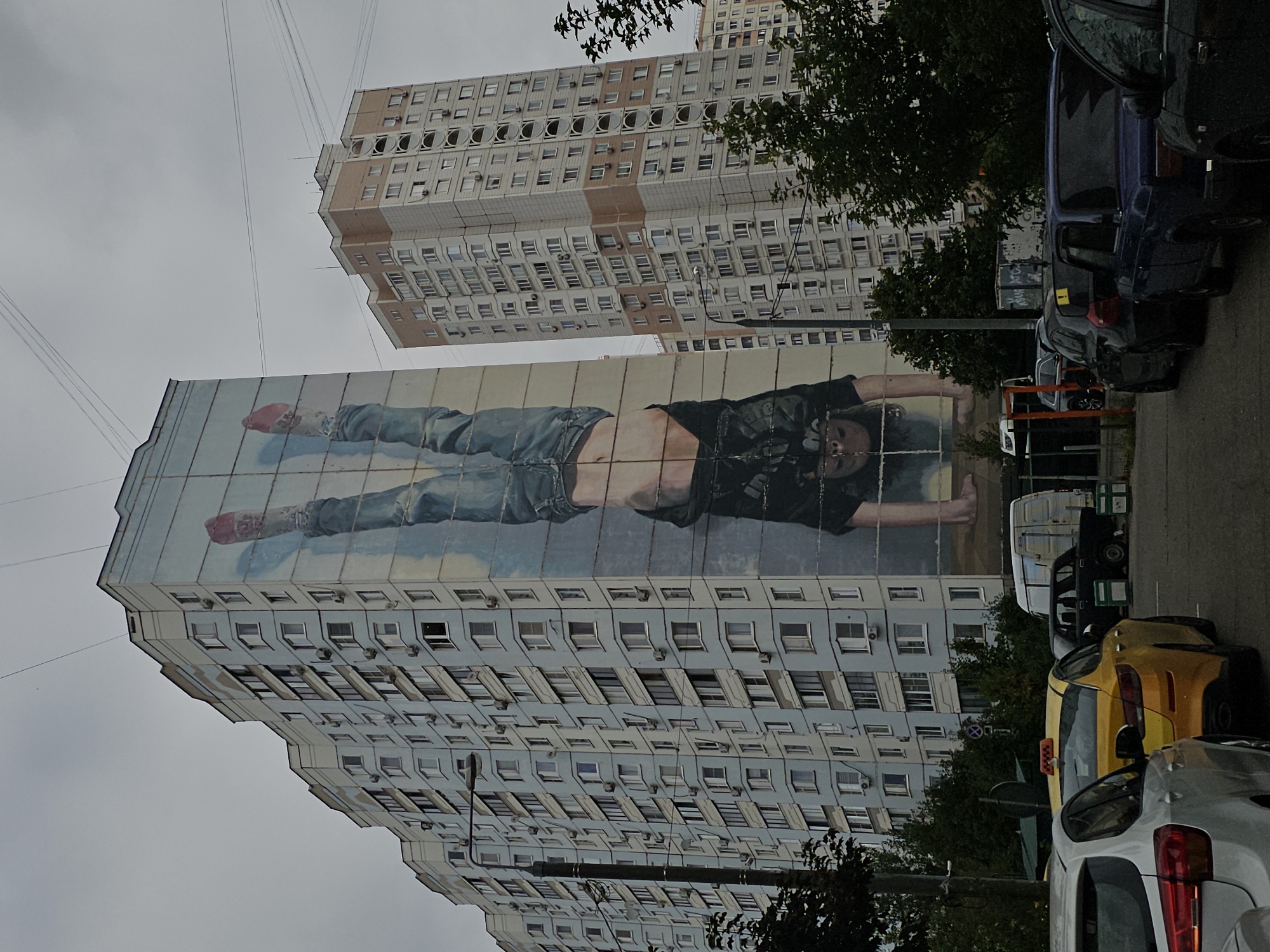 The image depicts a mural on the side of a light blue apartment building in Odintsovo, Russia. The mural shows a person in mid-handstand, upside down, wearing blue jeans and a dark patterned shirt. The person has red shoes on their feet. The building has many windows and air conditioning units. To the right of this building is a taller, cream-colored apartment building with some orange accents. In the foreground, parked cars are visible, including a yellow taxi with a "TAXI" sign on top. The sky is overcast. Trees and streetlights are also present in the scene.