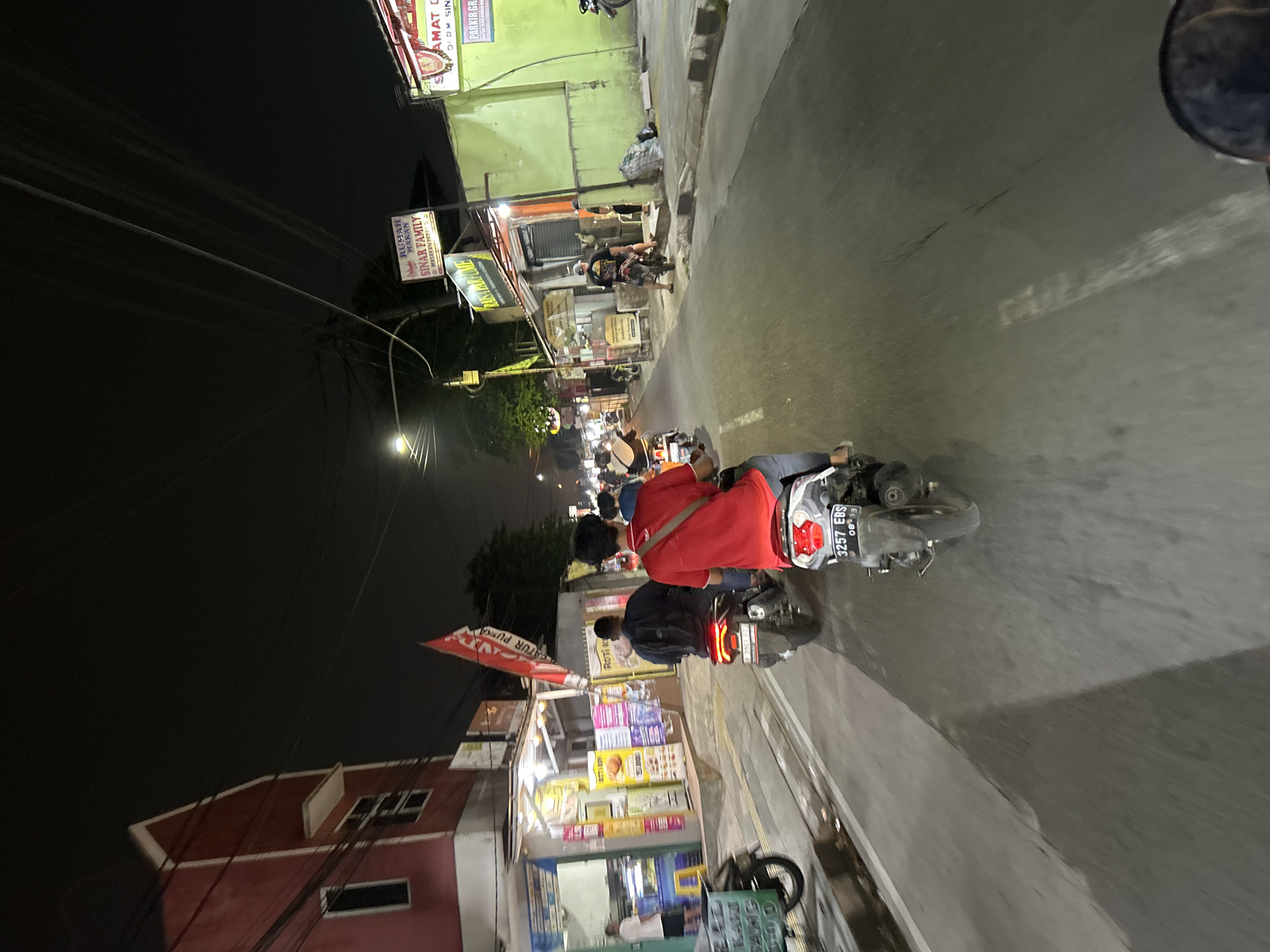This is a nighttime, slightly tilted, low-angle shot of a street scene in Depok, Indonesia. The image is dominated by a street lined with small businesses and illuminated by artificial lights.
In the foreground, two people are riding on a motorcycle, with the rider in front wearing a red shirt and a backpack. Behind them, another person rides a motorcycle. The motorcycle in the foreground has a license plate reading "3257 EBS 08-19". The road appears to be asphalt and is marked with a white line.
To the left of the road, there are several storefronts. One prominent sign reads "RUMAH MAKAN SINAR FAMILY" (Sinar Family Restaurant) with "MASAKAN PADIS" (Spicy Cuisine) below it. Another sign says "RAISA PARFUME". Further down the street, there are more illuminated signs and displays for various shops, including what appears to be a food stall with colorful products. In the background, more people and motorcycles are visible, suggesting a busy street at night.
To the far left, the corner of a red building with white trim is visible. Overhead, a network of power lines crisscrosses the sky, further emphasizing the urban setting. The overall atmosphere suggests a bustling commercial street at night, likely a popular spot for food and shopping. The lighting creates a vibrant, albeit somewhat dark, ambiance.