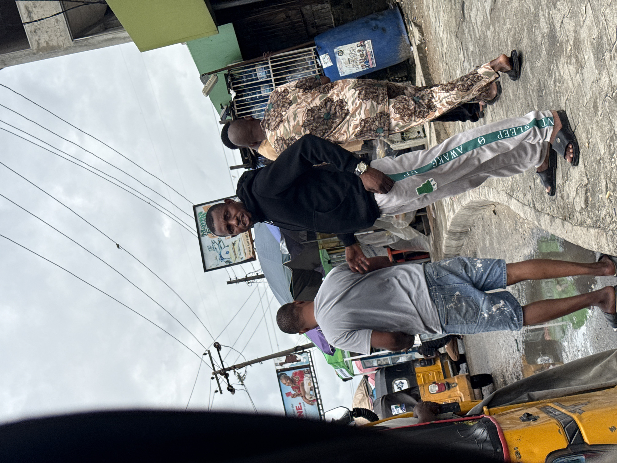 This is a medium shot taken outdoors on a cloudy day in Ado, Nigeria. The scene appears to be a busy street market or roadside area.
In the foreground and left of center, a yellow auto-rickshaw, also known as a "Keke," is partially visible, with its front and side showing. A person's dark-clad arm and shoulder are in the extreme left foreground, obscuring the view of the Keke's interior.
To the right of the Keke, a man wearing a grey t-shirt and distressed denim shorts stands facing away from the camera. He is wearing black slip-on sandals. His shorts have white paint splatters on them.
In the center of the frame, a man wearing a black hoodie and light grey sweatpants with a green stripe down the leg is looking directly at the camera. He is wearing a watch on his left wrist and black slip-on sandals. The green stripe on his pants has text that reads "AWAKE" and "SLEEP" vertically.
Behind these two men, another man is visible wearing a patterned short-sleeved dashiki in shades of brown and beige with dark brown floral designs. He is walking away from the camera.
Further in the background, there are several market stalls with umbrellas providing shade. One stall has a large blue barrel with signage. In the upper background, there are power lines and utility poles. Two large billboards are mounted on poles. One billboard depicts a woman holding a phone and the text "NE." The other billboard shows a close-up of a man's face and text, including "Mama F."
The ground is paved with interlocking stones and appears wet in places, suggesting recent rain. The overall impression is that of a vibrant, active street scene in an urban Nigerian environment.
