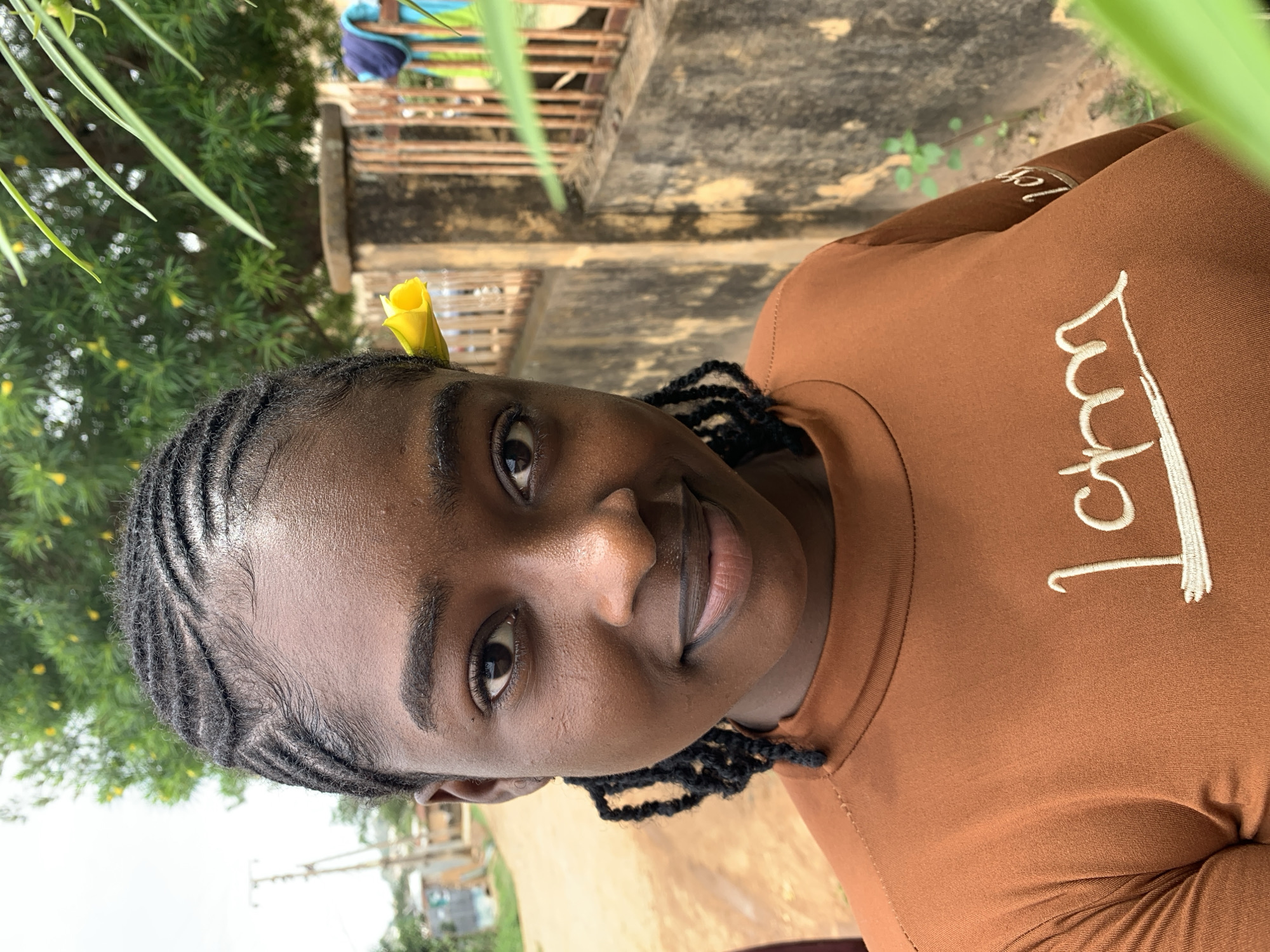 This image is a head-and-shoulders portrait of a young Black female, taken outdoors in the city of Rafin Par, Nigeria. She faces the viewer directly, exhibiting a subtle smile. Her dark hair is styled in intricate cornrows on the scalp, transitioning into loose braids that frame her face. A bright yellow flower is tucked behind her right ear. She wears a rust-brown, long-sleeved top with a mock neck, featuring white cursive embroidery on the chest that reads "I am."
The background on the left reveals a dirt road or path, with an out-of-focus utility pole and distant buildings under a bright sky. On the right, a weathered concrete wall with vertical metal bars forms a fence. Behind the fence, an out-of-focus tree with green leaves and numerous small yellow flowers, visually consistent with the flower worn by the subject, is visible. Beyond the fence, faint blue and turquoise fabrics are discernible, suggestive of drying laundry. The scene is illuminated by natural, diffused daylight.
