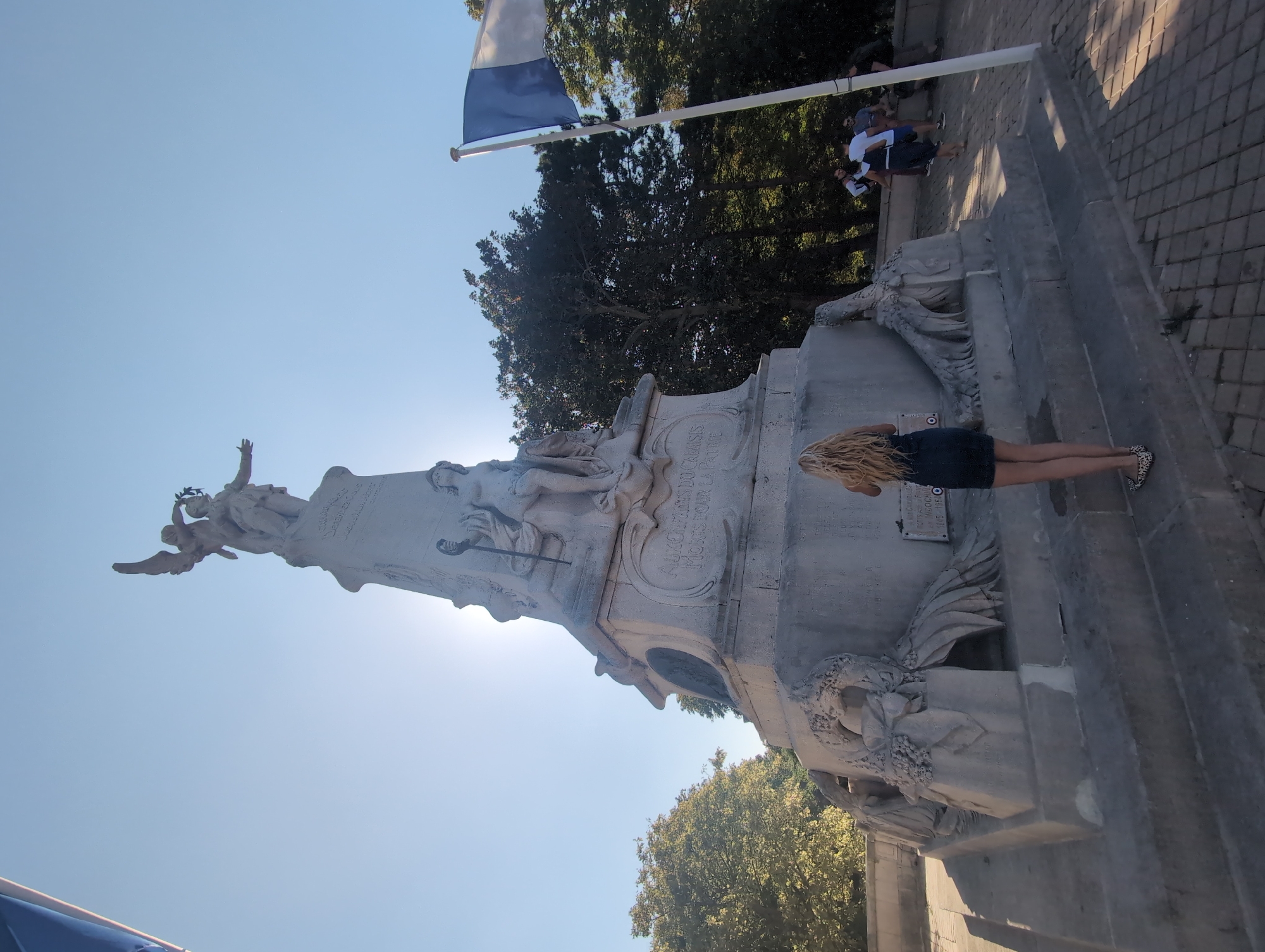 In a sunlit outdoor scene in Calais, France, a tall, ornate stone monument dominates the center of the frame. The monument, likely a war memorial, features a central statue of a winged figure crowning another figure, with additional sculptural elements and inscriptions on its base. To the right of the monument, a French flag flies on a flagpole. A woman with long blonde hair, wearing a dark blue dress and leopard-print flats, stands with her back to the camera, facing the monument. Several other individuals are visible in the background, some walking and conversing near the monument's base and a paved pathway. Lush green trees form a backdrop behind the monument and flag, suggesting a park or public square setting. The sky is clear and bright blue. The monument bears an inscription in French, "À UX ENFANTS DU CALAISIS MORTS POUR LA PATRIE," translating to "To the children of Calais who died for the homeland." Another plaque below this reads "INDOCHINE 1945-1954."