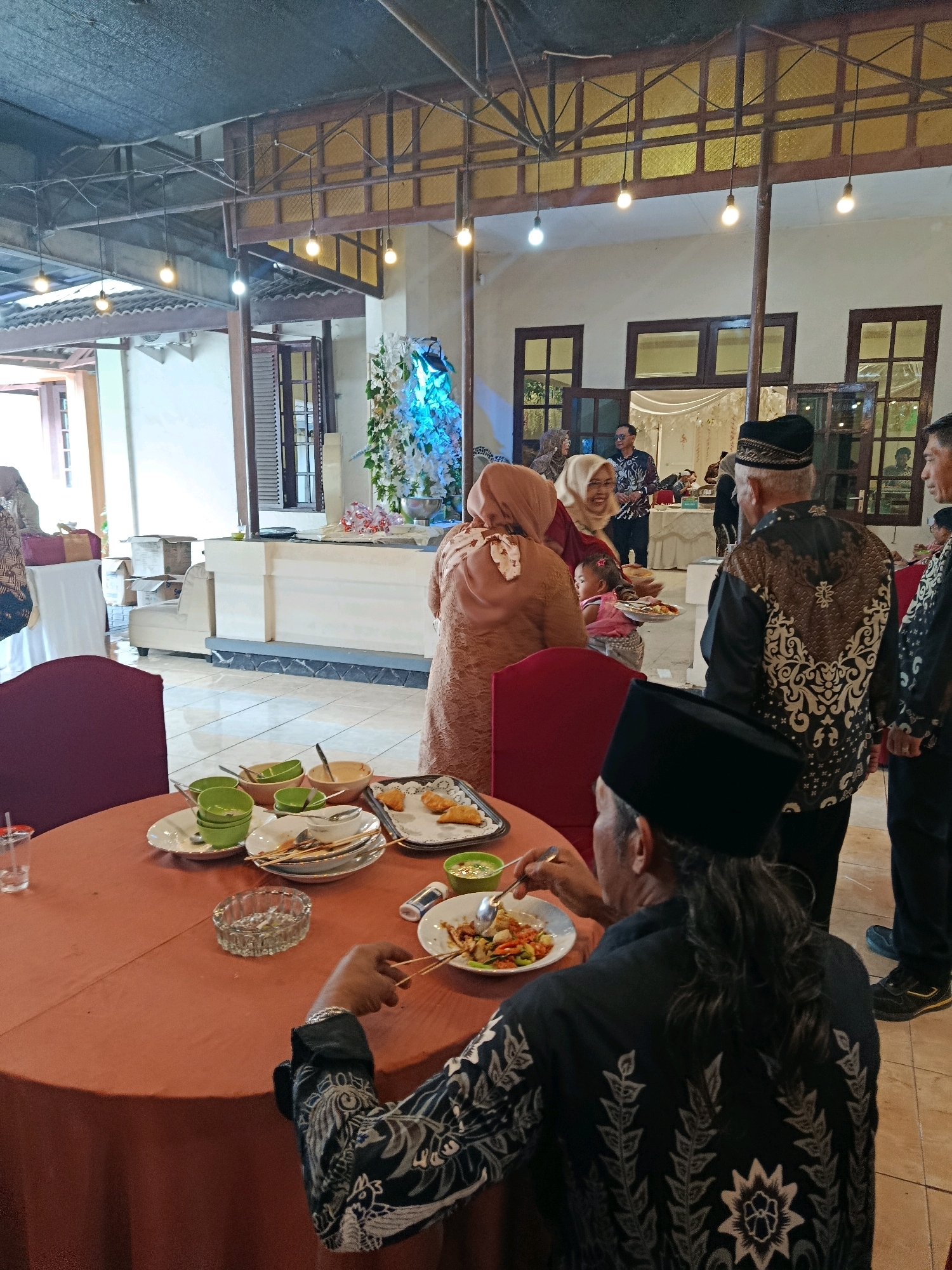The image depicts a gathering of people in what appears to be an indoor dining area, possibly a reception hall or restaurant, located in Tanggung, Indonesia. The environment is decorated with floral arrangements and ambient lighting from hanging bulbs and wall fixtures.
In the foreground, a round table is set with plates, bowls, and cutlery, suggesting a meal is in progress or has recently concluded. Several individuals are visible around the table and in the background. One man in the immediate foreground, facing away from the camera, wears a black cap and a black shirt with white embroidered patterns, and he is actively eating from a plate with a fork and skewers.
In the middle ground, a group of people are interacting. Some are dressed in traditional attire, including headscarves and batik shirts. One woman is holding a baby. Another man in a black and white patterned shirt stands with his back to the camera, also appearing to be engaged in conversation or observing the proceedings. In the background, additional tables and a buffet-style serving area with more floral decorations are visible. The overall atmosphere suggests a social event, possibly a wedding reception or a celebratory meal. The flooring is tiled, and the walls are painted in light colors. Wooden window frames and doors are visible throughout the space.