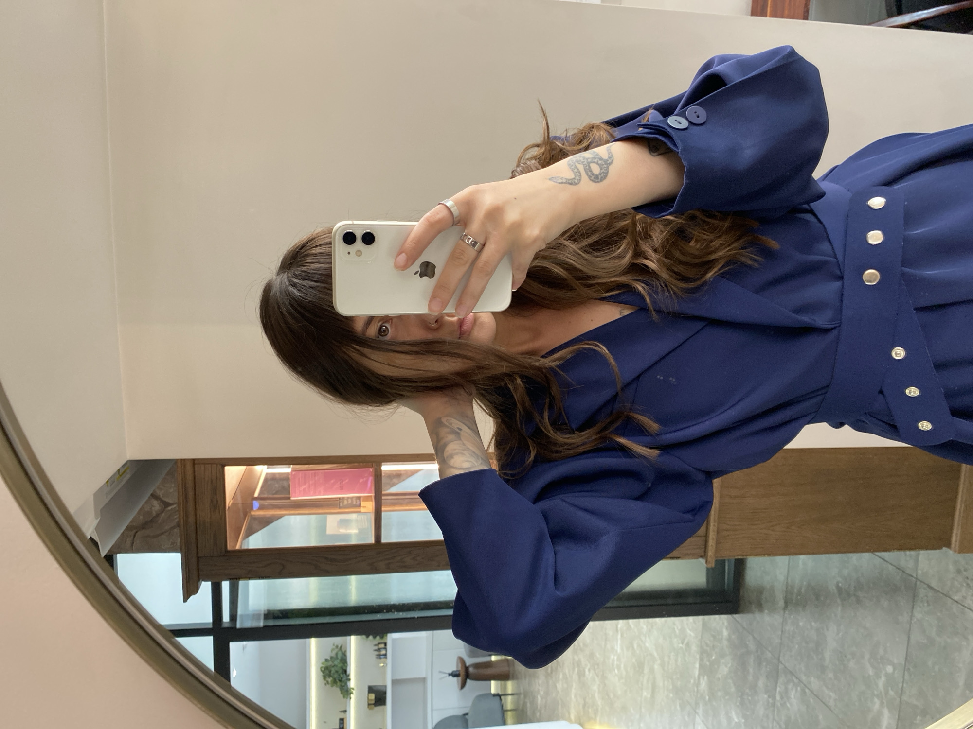 A young woman is taking a mirror selfie in a room with warm, neutral-colored walls. She has long, wavy brown hair and is wearing a dark blue blazer dress with a cinched waist and gold buttons. She has a tattoo of a snake on her forearm and is wearing two silver rings on her right hand. She is holding a white iPhone in her right hand, obscuring part of her face. In the background, a large, arched mirror reflects a room with a wooden cabinet filled with books and decorative items, as well as a glimpse of a plant on a shelf and furniture. The floor appears to be tiled. There is no specific information or visual cues to indicate the location is Votkinsk, Russia.