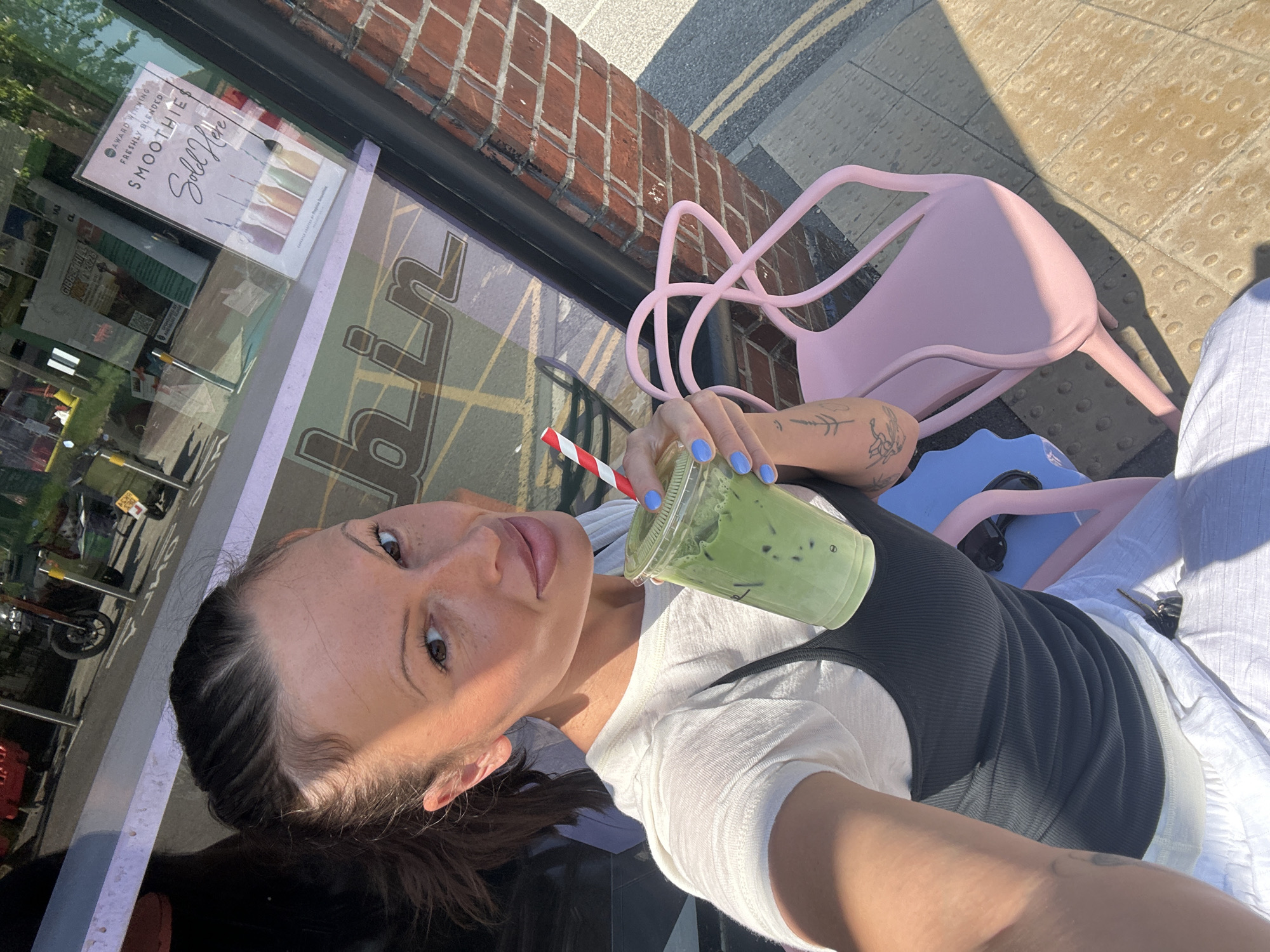 A self-portrait captures a woman sitting outdoors on a bright, sunny day, holding a green beverage. The woman, with dark hair and dark eyes, looks directly at the camera, exhibiting a calm expression. She wears a white short-sleeved top beneath a dark ribbed tank top. Her fingernails are painted a light blue, and she has visible tattoos on her left arm and shoulder. She holds a clear plastic cup containing a vibrant green, ice-filled drink, likely a matcha latte or smoothie, with a red and white striped straw.
The setting appears to be an outdoor seating area of a cafe or shop. A light pink, modern-style metal chair is positioned beside her, with a blue object partially visible further down. The ground is a light-colored paved surface, featuring tactile warning strips. In the background, a brick building facade is visible with a large window. Reflections in the window reveal an outdoor street scene, including greenery, what appears to be parked vehicles, and other urban elements.
Several pieces of text are visible on a sign displayed in the shop window: "AWARD WINNING FRESHLY BLENDED SMOOTHIE" and "Sold Here", accompanied by illustrations of various smoothies. Part of what appears to be the shop's name, "biin", is also reflected in the window. Another reflected poster shows text that includes "GHOST HILL 1-2023". Strong sunlight casts distinct shadows, indicating it is daytime. The image conveys a relaxed moment at an outdoor establishment in Waverley, United Kingdom.