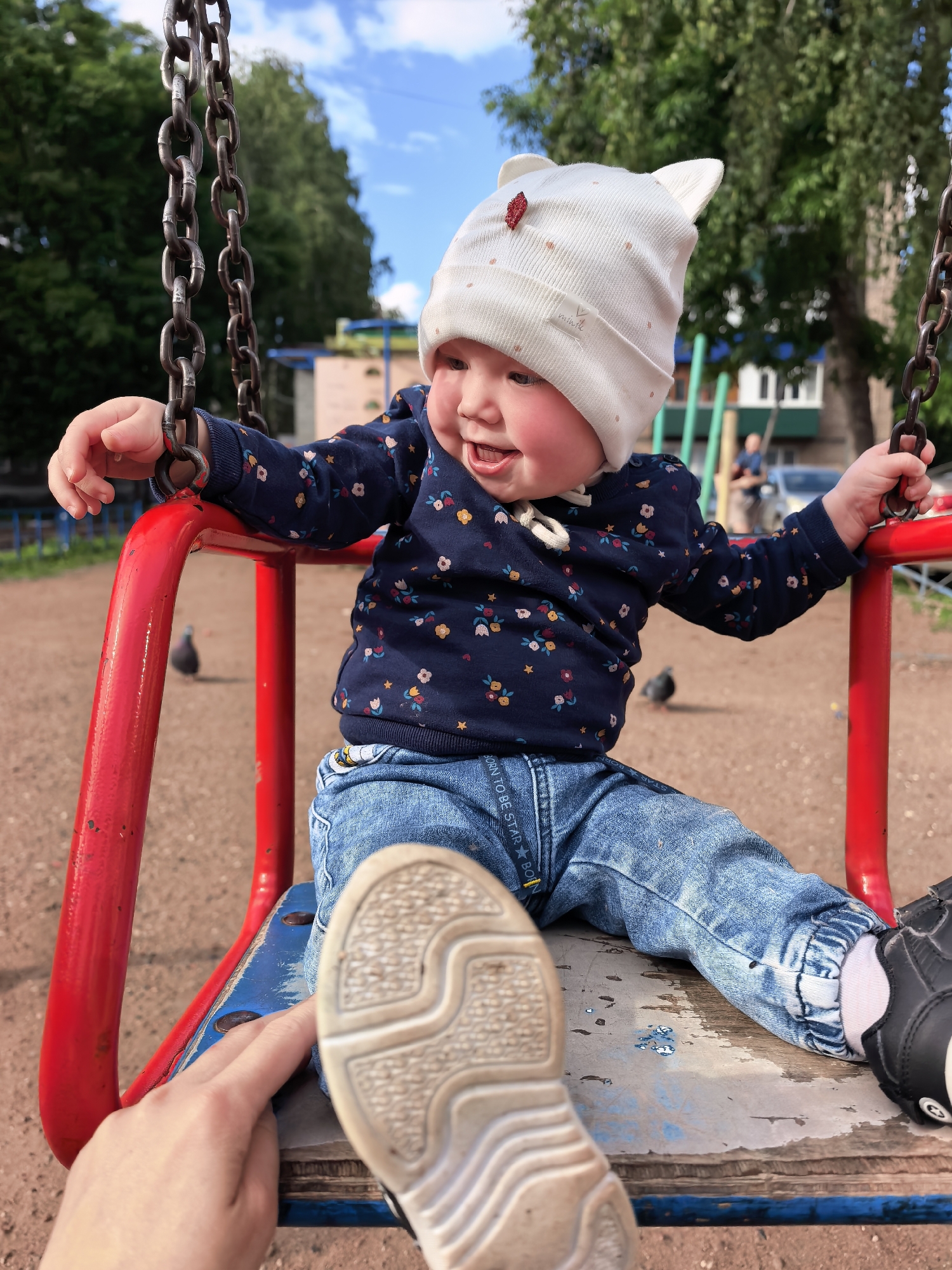 A young child, approximately six to eighteen months old, is seated on a red-framed swing with a blue-painted wooden or metal seat. The child wears a white knitted hat with ear-like protrusions and a small red embellishment, a dark blue long-sleeved shirt with a multicolored floral pattern, and light blue denim jeans with elasticized ankles. Black velcro-closure shoes are on the child's feet. The child's left hand grasps one of the metal chains of the swing, and the face shows an open-mouthed expression of enjoyment, with rosy cheeks. An adult's right hand is visible in the lower left foreground, touching the child's left foot, which wears a beige-soled shoe. The background features a dirt or sand playground surface. Two pigeons are visible on the ground behind the child. Beyond the playground, green trees dominate the midground, with a multi-story residential building, likely an apartment block, partially visible to the right. A person in a blue shirt is faintly discernible near the building. The sky is partially cloudy blue. The scene is located in a playground within the city of Sterlitamak, Russia.