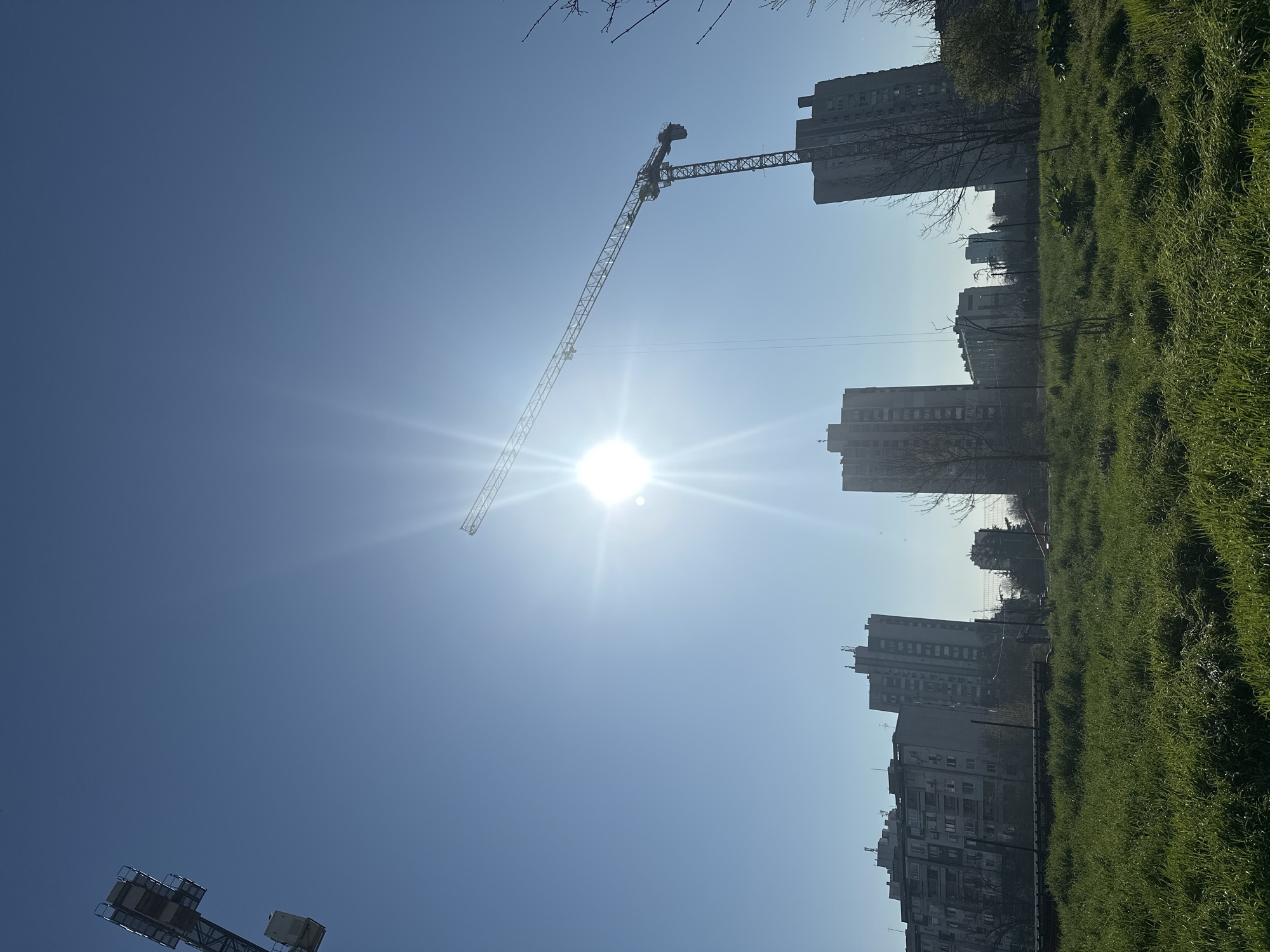 The image presents a view of an urban area. In the foreground, there's a patch of green grass, likely a park or open space. In the background, there are several high-rise buildings, suggesting a residential or commercial area within a city. A construction crane is visible against the bright sky, possibly indicating ongoing construction or development in the area. The weather appears to be sunny, as indicated by the bright blue sky and the intense sunlight that dominates the view. The city location is Unknown, Unknown.