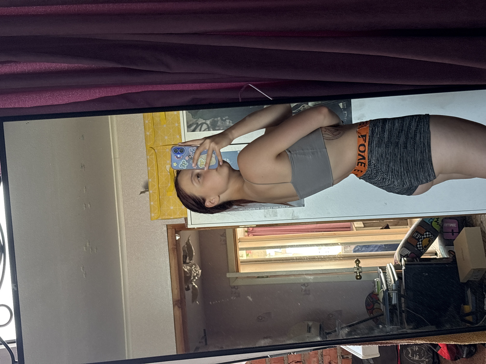 A woman is taking a selfie in a mirror. She is wearing a grey sports bra and grey athletic shorts with an orange waistband that reads "FOREVER." Her phone has a colorful case. The reflection in the mirror reveals a cluttered room with a window. The location is unknown; Прикамский is not mentioned or otherwise discernible from the image.