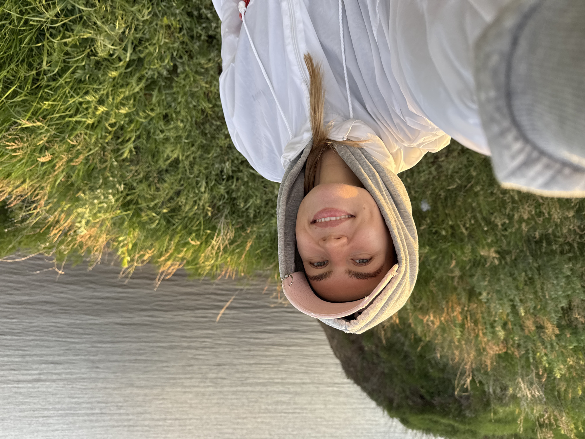 A young adult female is depicted in a close-up, waist-up selfie taken outdoors in the city of Unknown, Unknown. She looks directly at the camera with a broad smile, revealing her teeth. Her blue eyes are open, and her eyebrows are well-defined. She wears a grey hooded sweatshirt underneath a white, lightweight, zippered jacket with drawstrings. A pink baseball cap with a small, silver-colored hoop piercing on its brim is visible beneath the hood. Her light-colored hair is partially visible on her right shoulder. The background features a steep, grassy slope with various green grasses, some with golden-brown tips illuminated by warm, directional sunlight. Beyond the slope, a large body of calm, rippled water extends to the horizon. The scene is lit by golden hour sunlight, casting warm tones across the subject's face and the landscape.
