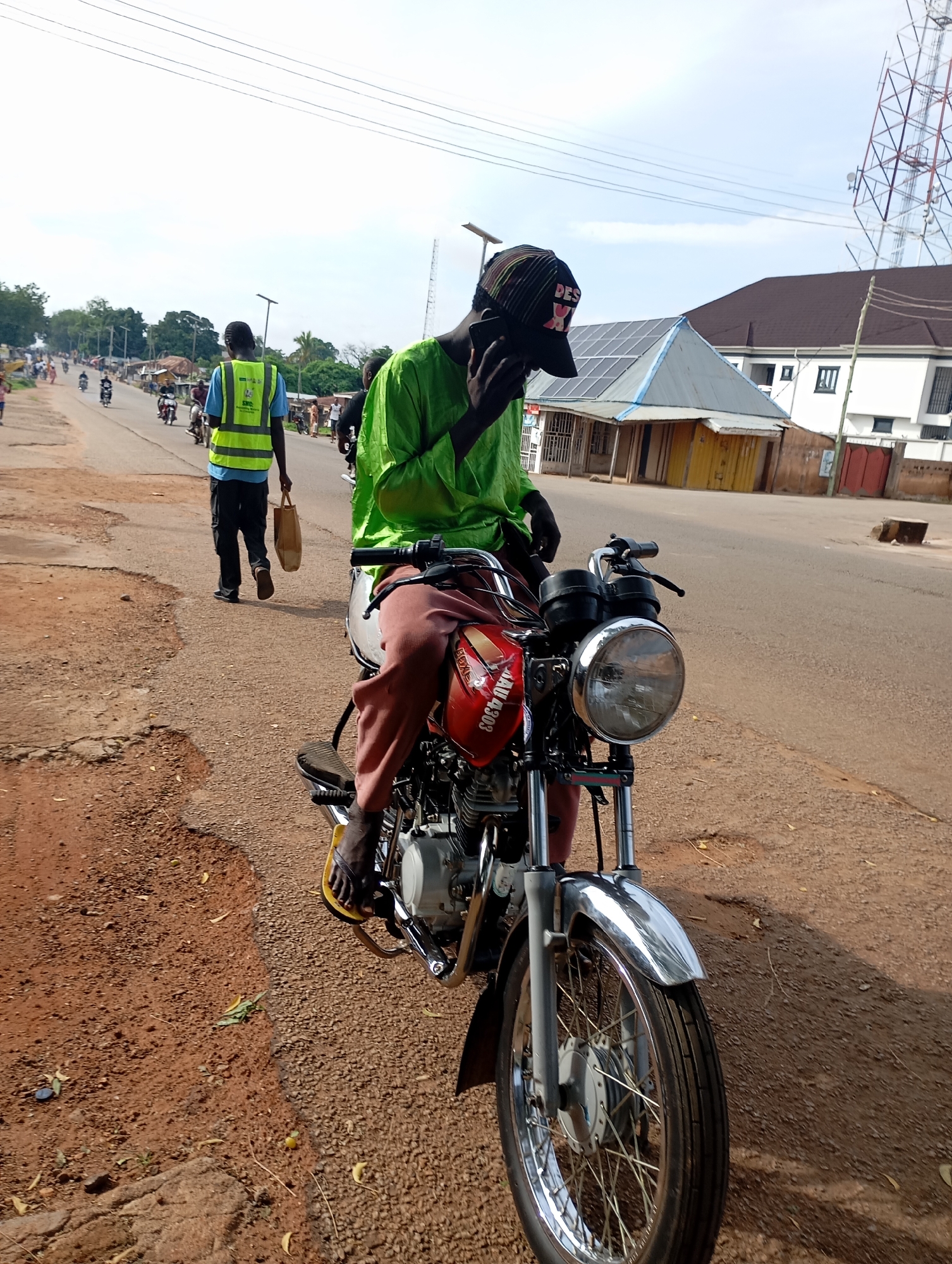 In the city of Shendam, Nigeria, an individual wearing a bright green outfit and a black cap with the word "DES" visible is seated on a red motorcycle. They are holding a black mobile phone to their ear, appearing to be engaged in a conversation. The motorcycle is parked on the side of a paved road, with loose dirt and gravel on the shoulder. In the background, other people are walking and riding motorcycles, and a man wearing a reflective yellow vest is walking away from the camera carrying a brown paper bag. Buildings with solar panels on their roofs and a tall telecommunications tower are also visible in the distance. The scene depicts a typical street view in an urban setting in Nigeria.