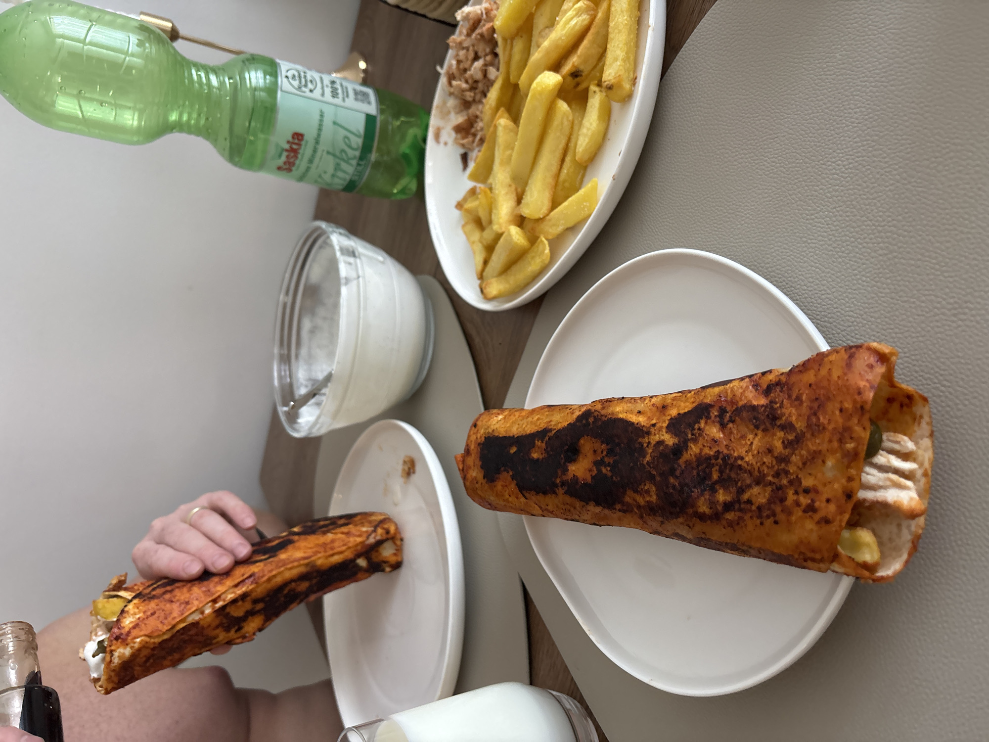A close-up, eye-level shot shows a meal at a table, likely in Viernheim, Germany. In the foreground, a person's hands are visible, one holding a large, grilled wrap with a reddish-orange exterior, and the other holding a dark liquid in a small bottle, preparing to pour it over the wrap. The wrap is filled with visible chicken, pickles, and a white sauce. Another identical wrap rests on a white plate to the left. To the right, a white platter holds a pile of golden-brown french fries and a portion of shredded meat, possibly also chicken. A clear glass of milk and a small bowl of white sauce are also on the table. A green bottle of "Saskia" mineral water stands in the background. The setting appears to be an indoor dining area, with a light-colored wall and a wooden table surface.