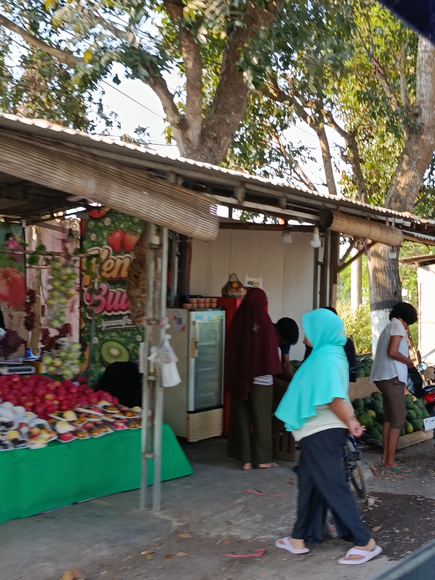 The image depicts an outdoor fruit stand in Ramban Wetan, Indonesia, under a canopy of large trees. The stand displays a variety of fruits, including apples and grapes, with some fruits packaged in plastic. Several individuals are present at the stand, some appearing to be customers and others possibly vendors. The scene is bustling with activity, suggesting it is a typical day at a local market.