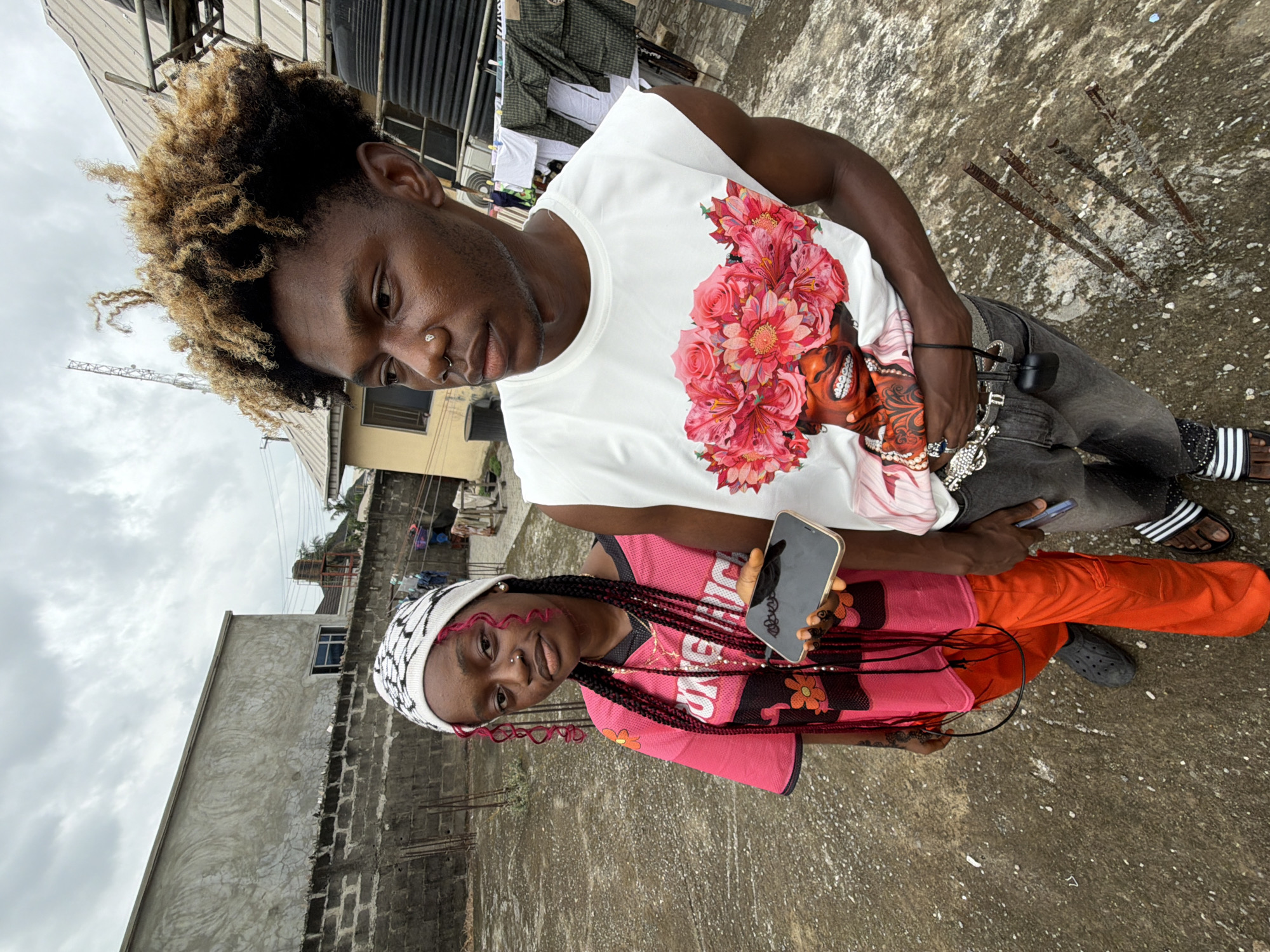 The image shows two people standing on what appears to be a rooftop in Ado, Nigeria. The man wears a white tank top with a floral design, gray pants, and striped sandals. He has dyed blonde hair. The woman wears a pink athletic top, orange pants, a white headscarf, and holds a smartphone. The background includes buildings, a cloudy sky, exposed metal rods, and clotheslines.