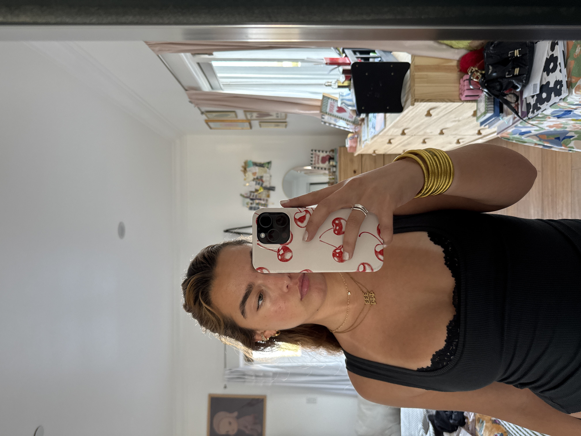A young woman is taking a selfie in a mirror. She is wearing a black tank top with lace trim, a gold necklace, and multiple gold bangles on her wrist. She is holding a white iPhone with a cherry-patterned case.
The background shows a room in a house. There's a window with sheer pink curtains, framed pictures on the wall, a dresser with various items on top including a black object that might be an electronic device, and various decorative items.
Based on the interior design and architectural style, the location is likely in a residential area of Los Angeles, United States of America.
