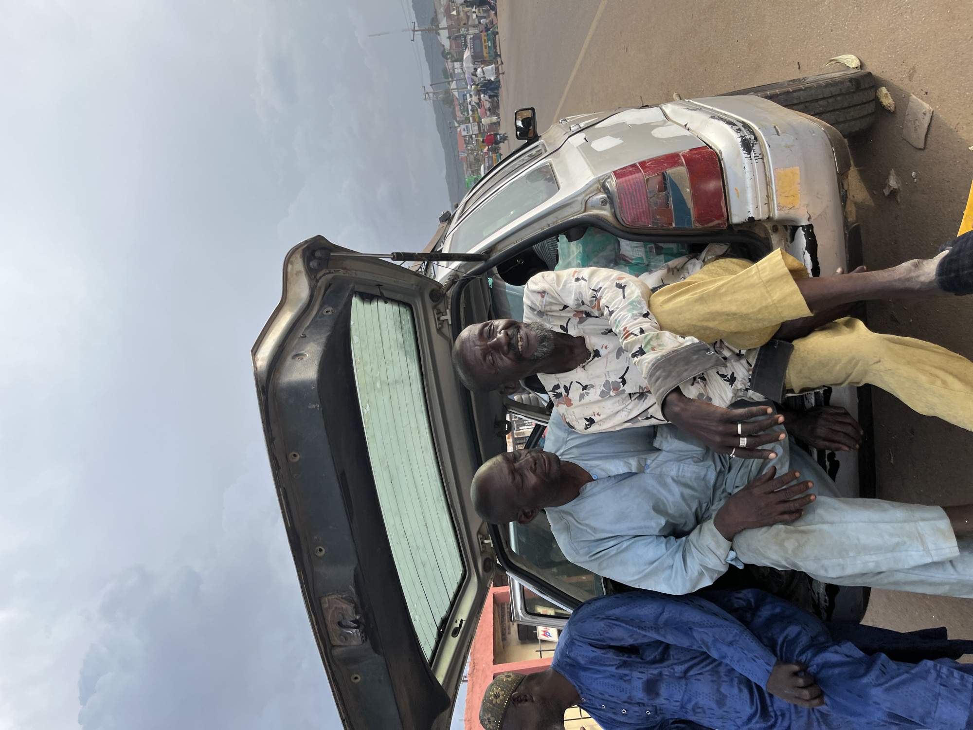 In Rafin Par, Nigeria, three men are seen gathered around the open trunk of a silver sedan. The trunk is open, revealing the interior of the car and the men are seated within or leaning against it. One man, in a white shirt with a floral pattern, is laughing heartily while seated in the trunk. He wears yellow pants and has a beard. Next to him, another man in a light blue traditional outfit is looking down, his arms crossed. A third man, dressed in a dark blue traditional outfit, is partially visible at the bottom of the frame, also appearing to be in conversation or resting.
The setting appears to be outdoors, with a dusty road surface visible and a backdrop of buildings and distant hills under a cloudy sky. The overall scene suggests a casual gathering or a moment of rest during travel. The weather appears overcast. The car, a sedan, shows signs of wear and tear, with some damage to its rear bumper and taillight. No visible text is present in the image. The time of day is not explicitly clear but the lighting suggests it is daytime. The emotions captured are primarily positive, with the laughing man conveying joy and amusement.