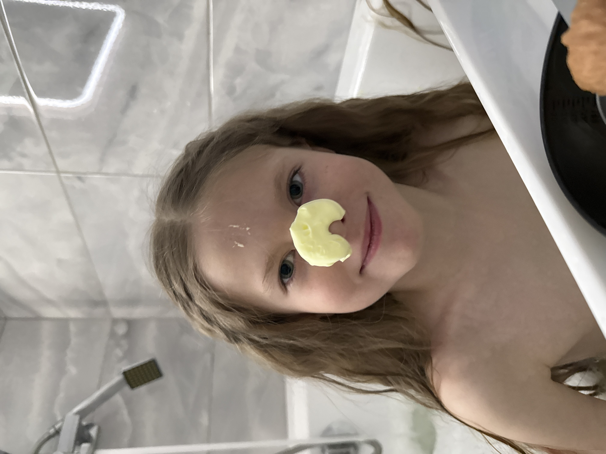 A young girl is in a bathroom. A blob of yellow substance is placed on her nose. She has fair skin, blue eyes, and long, light brown hair. A dab of the same yellow substance is also on her forehead. The background shows white and grey tiled walls and a showerhead. Part of a white bathtub and a black plate with food on it are also visible. The image was taken in a bathroom but there is no evidence of it being in the city of Izhevsk, Russia.