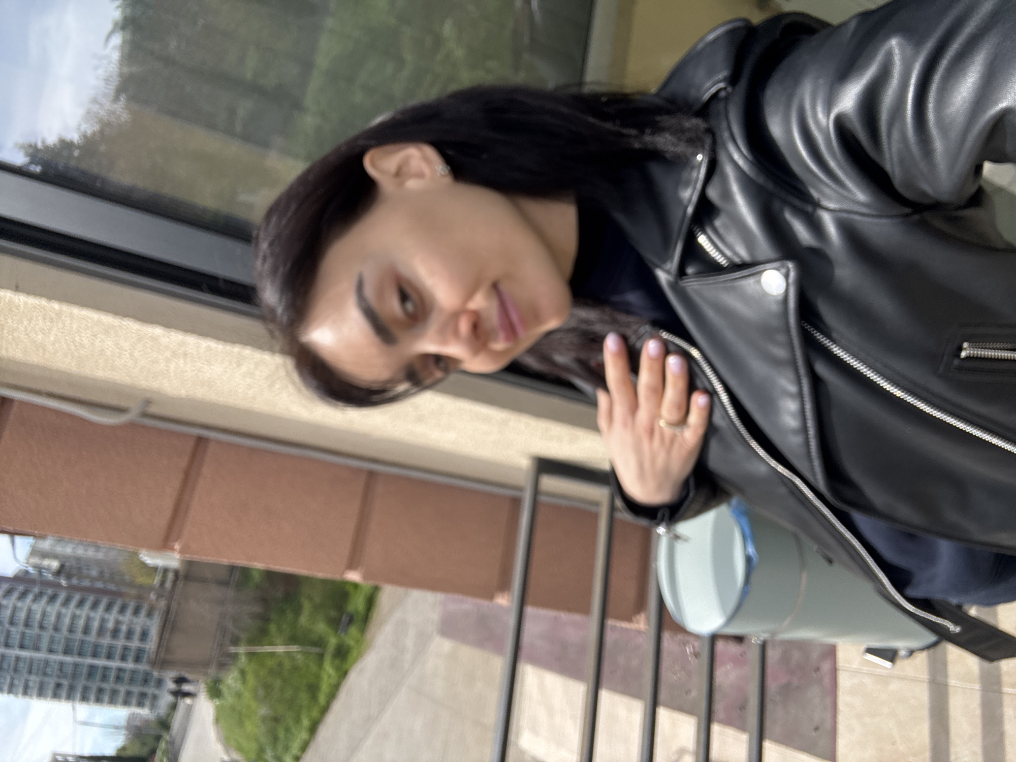 The image depicts a woman with dark hair wearing a black leather jacket. She is standing on a balcony with a railing, overlooking a cityscape. In the background, there are buildings, trees, and possibly a street or park. A cylindrical, light-colored object, possibly a container or bucket, is positioned near the balcony railing. The scene suggests an urban environment, likely in Tbilisi, Georgia, as indicated in the prompt.