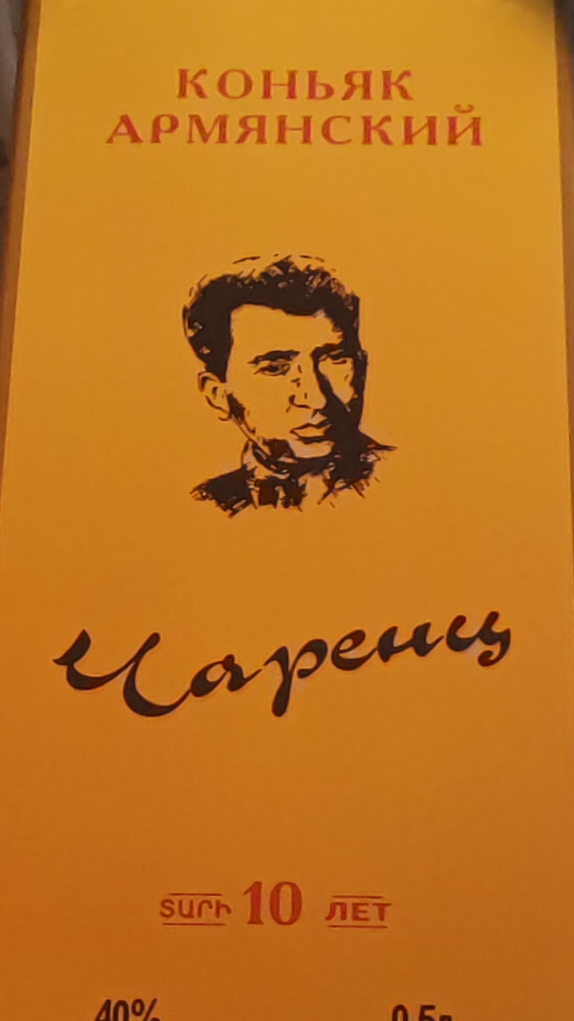 The image is a close-up of a product label with a warm orange background. At the top, in red Cyrillic text, it reads "КОНЬЯК АРМЯНСКИЙ" which translates to "Armenian Cognac". Below this text, a black silhouette of a man's head and shoulders is depicted, facing to the right with a stern expression. Prominently displayed in the lower half of the image is a large, stylized signature in black cursive script that reads "Gapenus". Further down, in a smaller red font, are the numbers "10" followed by "ЛЕТ" (years) and the Armenian word "ՏԱՐԻ" (years) which appears to be a misspelling or alternative script for "տարի". Below this, partially visible at the bottom left, is "40%" and at the bottom right, "0.5л" (0.5 liters). There is no visible indication of the city of Sochi, Russia, or any environmental context that would place it there.