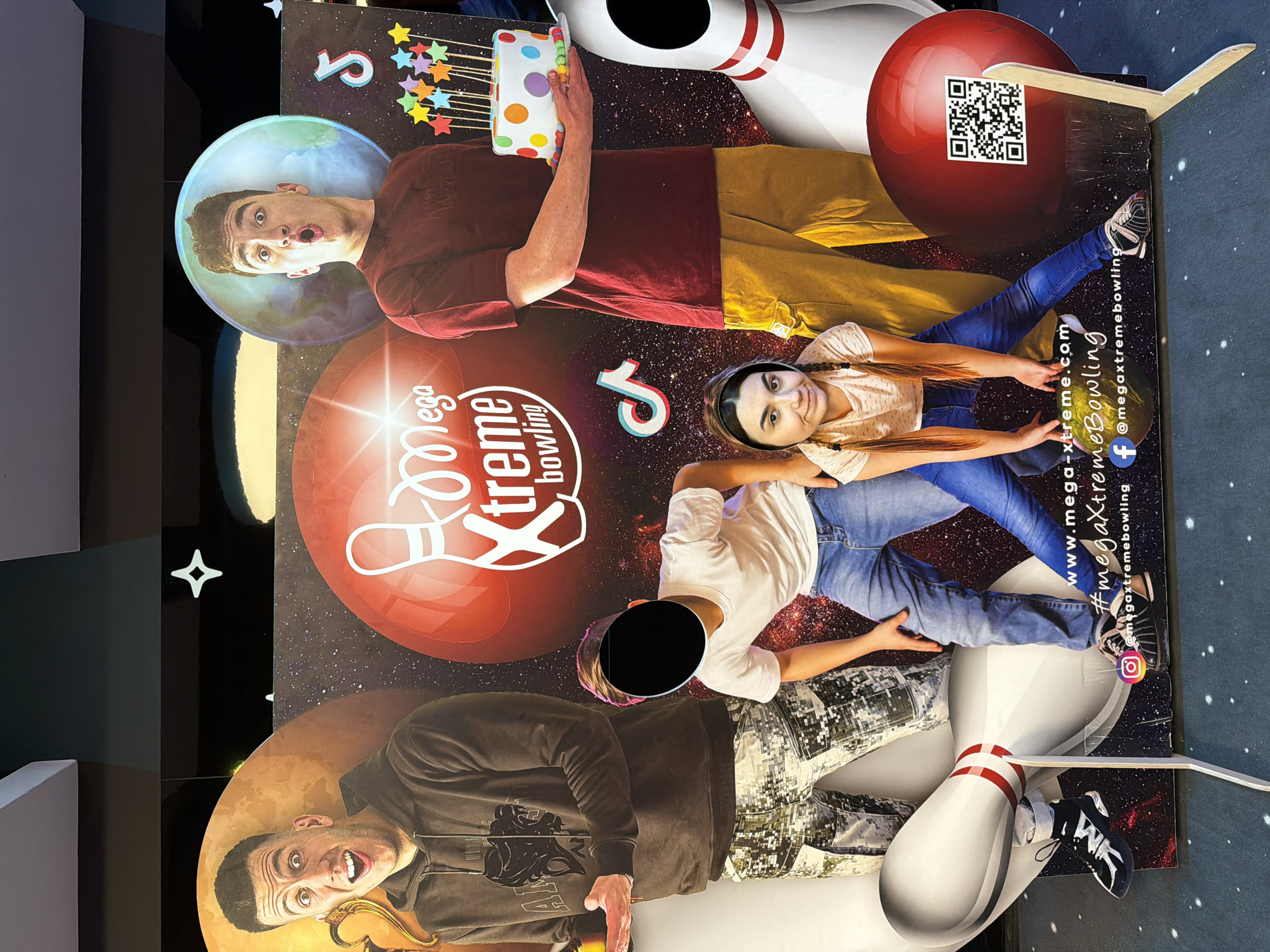 The image appears to be a poster for "Mega Xtreme Bowling," featuring several digitally inserted people posing as if interacting with bowling equipment. One person is in a planetary bubble, holding a birthday cake with candles, and positioned near a bowling pin. Another person is lying next to a bowling ball featuring a QR code. A third person is next to a bowling pin. The poster includes social media information for Mega Xtreme Bowling. The environment is unclear and there is no indication of the city of Sofia, Bulgaria.