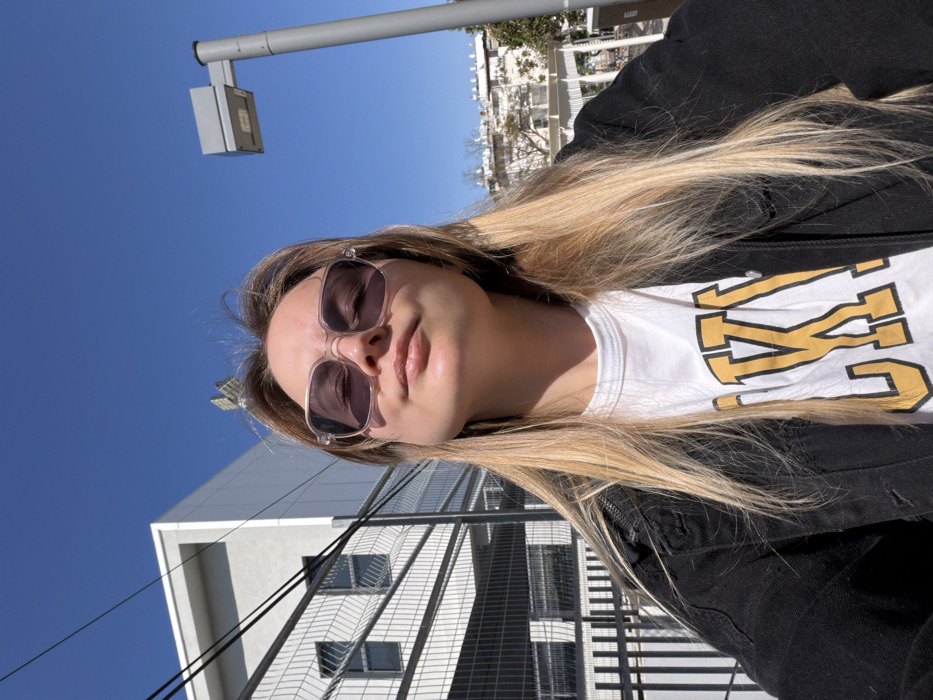 A young adult female, with long blonde hair, is centrally positioned in the foreground, looking upwards and towards the camera. She is wearing clear-framed sunglasses, a white t-shirt with yellow lettering (partially visible as "LUK!"), and a black denim jacket. Her eyes are closed or squinting against the bright sunlight, which illuminates her face from above. To her left, a dark metal mesh fence runs horizontally, behind which a multi-story, modern white building with dark-framed windows is visible. Several black utility wires are stretched across the clear blue sky above the fence and building. To her right, a tall, grey streetlamp with a rectangular light fixture stands. In the further background, trees, a white pergola-like structure, and residential buildings are discernible under the clear blue sky. The scene is brightly lit by direct sunlight on a clear day. Based on the modern architecture, urban infrastructure, and landscaped public spaces, the location is identified as the **Sarona complex in Tel Aviv, Israel**.