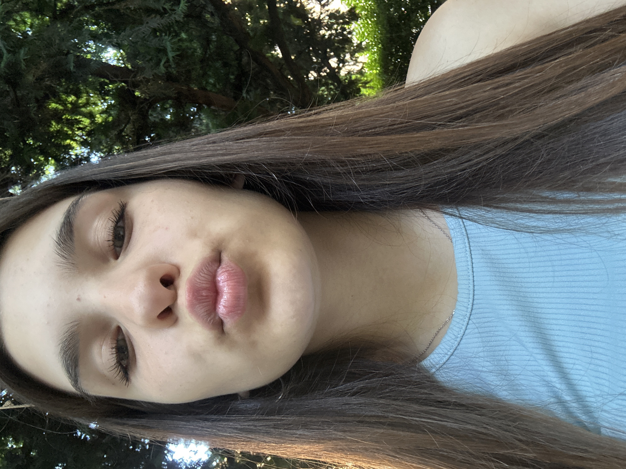 A young woman with long brown hair and light blue ribbed tank top poses for a close-up selfie outdoors. Her lips are puckered, and her eyes are looking towards the camera. The background consists of lush green foliage and trees, with sunlight filtering through the leaves. The overall impression is that of a casual, candid moment captured in a natural setting, possibly a park or garden in Shumen, Bulgaria.