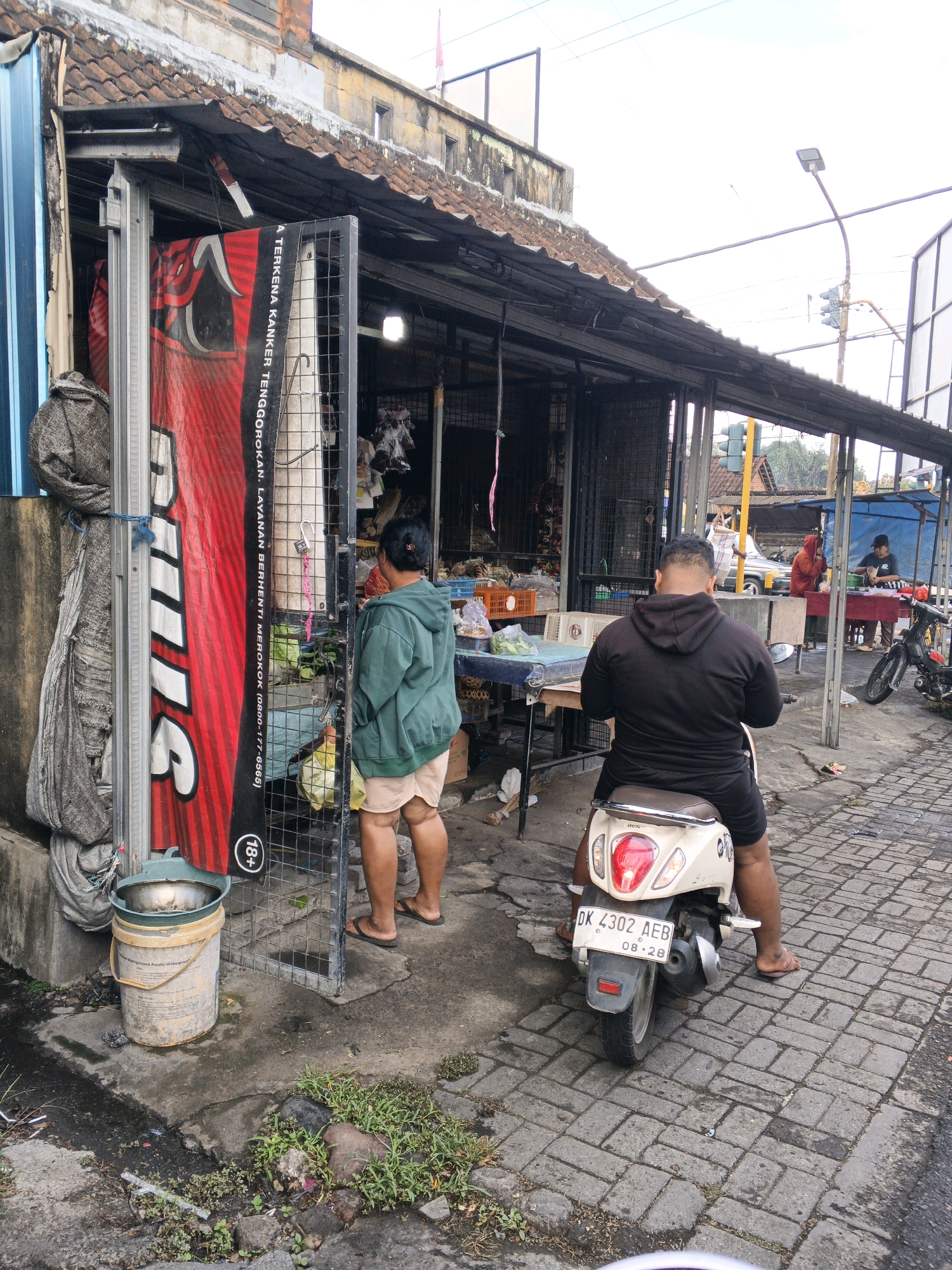 In Denpasar, Indonesia, a street scene unfolds with a small market stall in the foreground. A person in a green hooded jacket and light-colored shorts stands near a metal gate, looking into the stall. To the right, a man in a black hoodie and shorts sits on a beige scooter, facing away from the camera. The scooter has a license plate that reads "DK 4302 AEB" and "08-28". Behind the stall, other market activities are visible, with people interacting and goods displayed on tables. The ground is paved with interlocking bricks, and a large red banner with white text is prominently displayed on the left side of the stall. A metal bucket with a metal basin inside sits on the ground near the gate. The overall environment appears to be an outdoor marketplace on a cloudy day.
