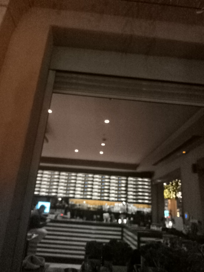 The image captures a dimly lit indoor scene, looking into a commercial establishment, likely a bar or restaurant, through a dark, architectural opening or doorway. The framing on the left and top suggests an archway or a thick wall structure.
Inside, the ceiling features several small, bright recessed lights, illuminating the space. A dominant feature is a large, grid-like wall display in the background, appearing to be a well-stocked wine or liquor rack with numerous bottles, many of which are illuminated from behind or below, creating a luminous pattern. Below this display is a bar counter, where various bottles and glassware are faintly visible.
On the left side of the bar, a single person is seen, partially obscured, appearing to be seated or standing. A bright blue screen, possibly from a laptop or tablet, is prominently visible in front of them. In the foreground, there are dark, possibly striped structures that could be banquettes, planters, or partitions. To the far right, some out-of-focus, warm-toned lights, resembling decorative string lights, add a festive touch.
The setting suggests evening or nighttime due to the reliance on artificial lighting. No specific weather conditions are discernible. There are no explicit location cues visible in the image itself to confirm Palaio Faliro, Greece, apart from the provided context. No emotions are discernible, and there is no visible text.