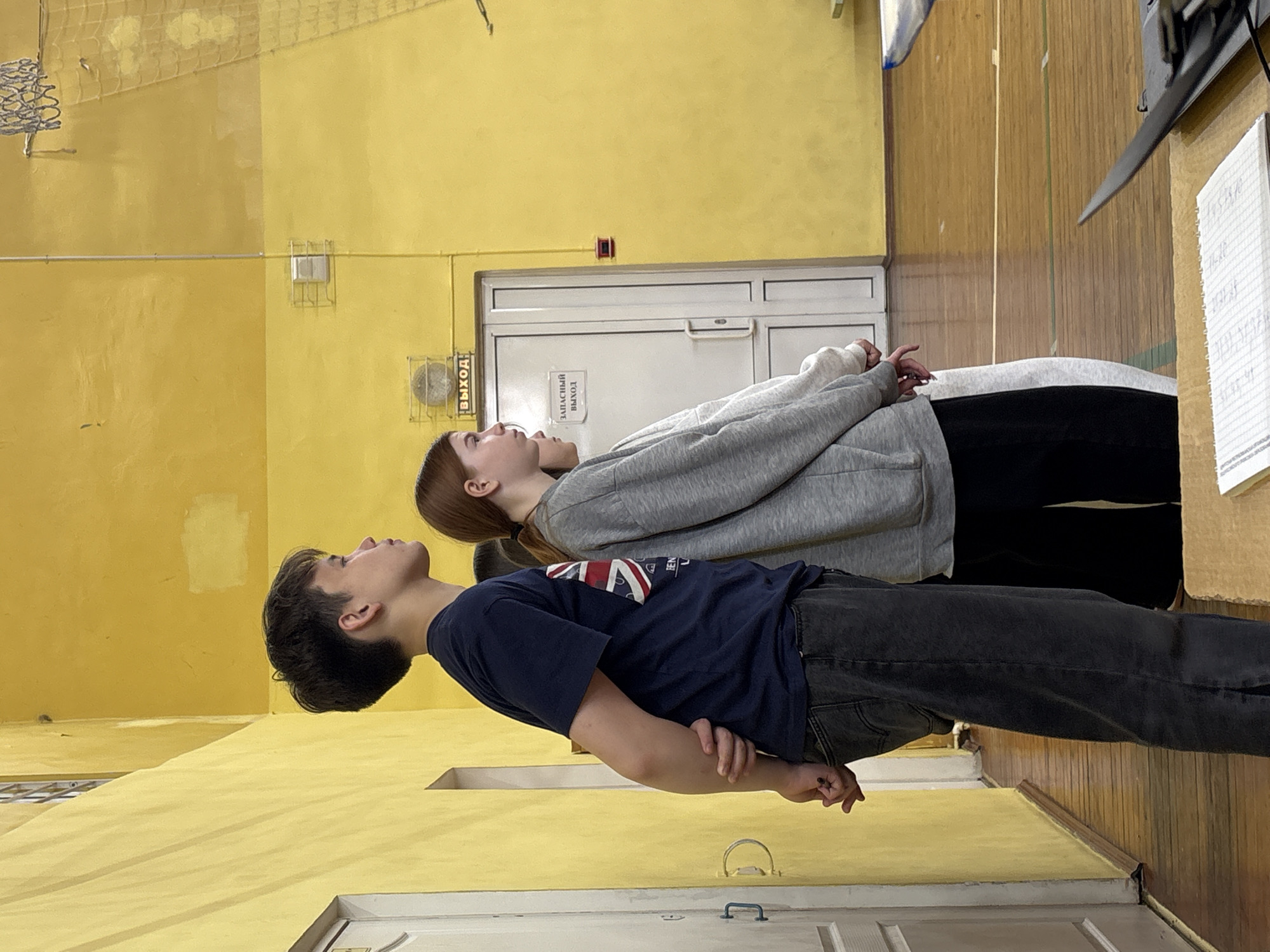 Two individuals, a male and a female, are standing in a gymnasium. The male, in the foreground, wears a dark blue t-shirt and grey jeans. The female, slightly behind him, is wearing a grey sweatshirt and dark pants. They both have their heads tilted back. The gymnasium has yellow walls. A basketball hoop is visible in the upper left corner. A door with Russian text "Выход" and "Запасный выход" (Exit and Emergency exit) is located behind the subjects. The location is Izhevsk, Russia.