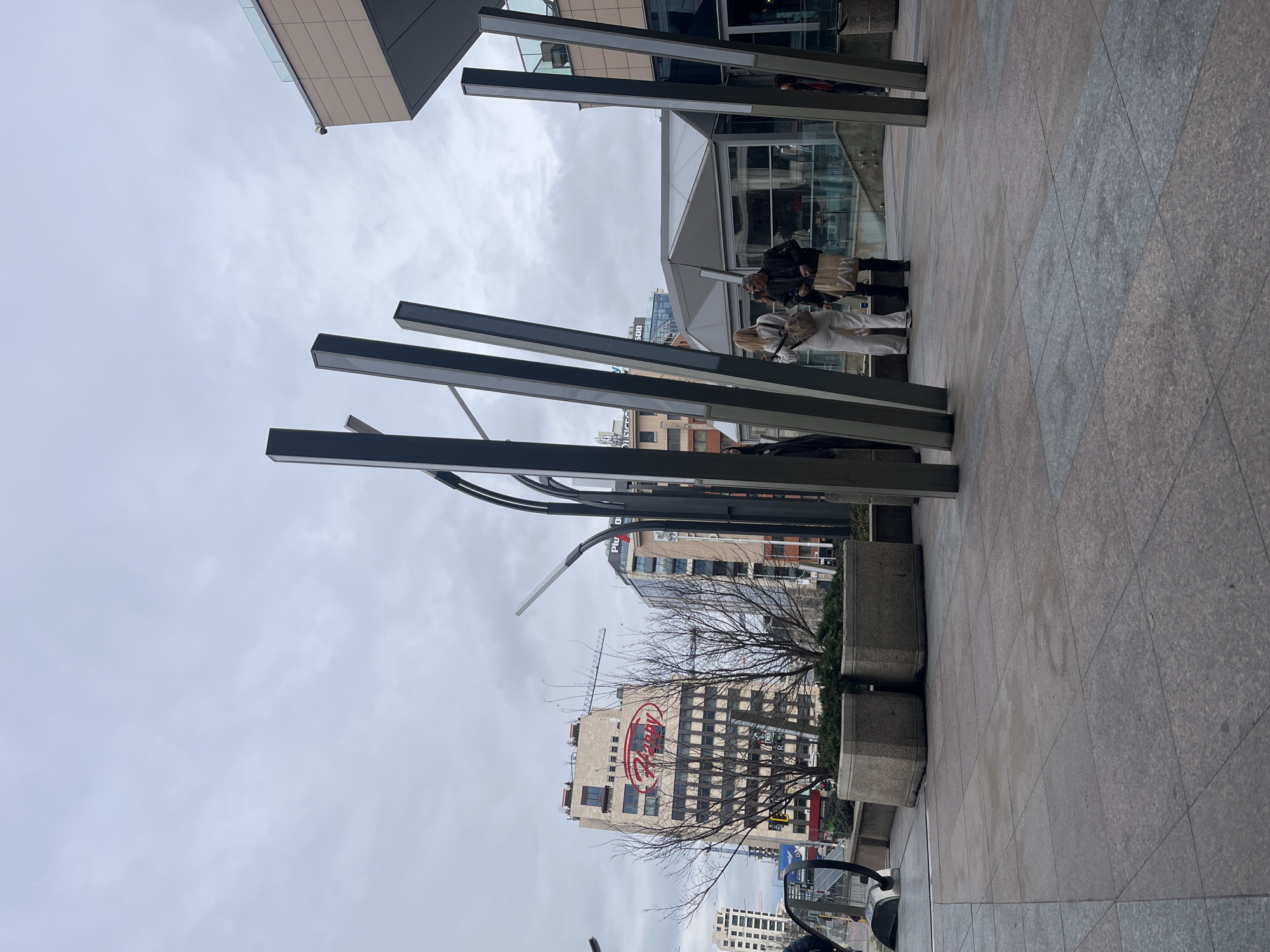 The image depicts an urban scene in Sofia, Bulgaria on an overcast day. In the foreground, there are several dark metal vertical architectural elements, partially obscuring a view of the city. Behind these structures, a group of people stand near the entrance of a modern building with glass walls. Further back, there is a building with the red "Happy" restaurant logo prominently displayed. A bare tree is positioned near the building. The ground is covered with gray tile. A portion of a vehicle is visible at the bottom of the frame.