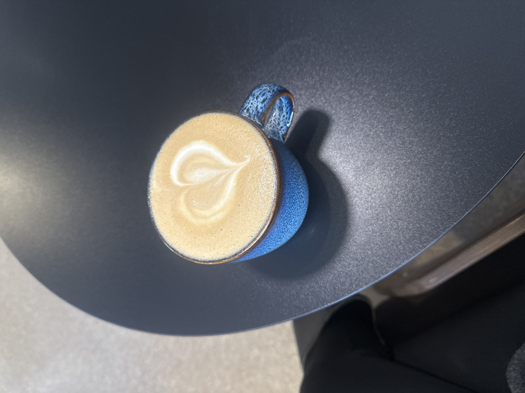 An overhead shot of a cappuccino with latte art in the shape of a heart, resting on a round, grey table. The cup is blue with a textured handle. Part of a chair is visible in the lower right corner. The location is unknown, and there is no information to suggest this photo was taken in Chaikovsky, Russia. There are no people depicted in the image.
