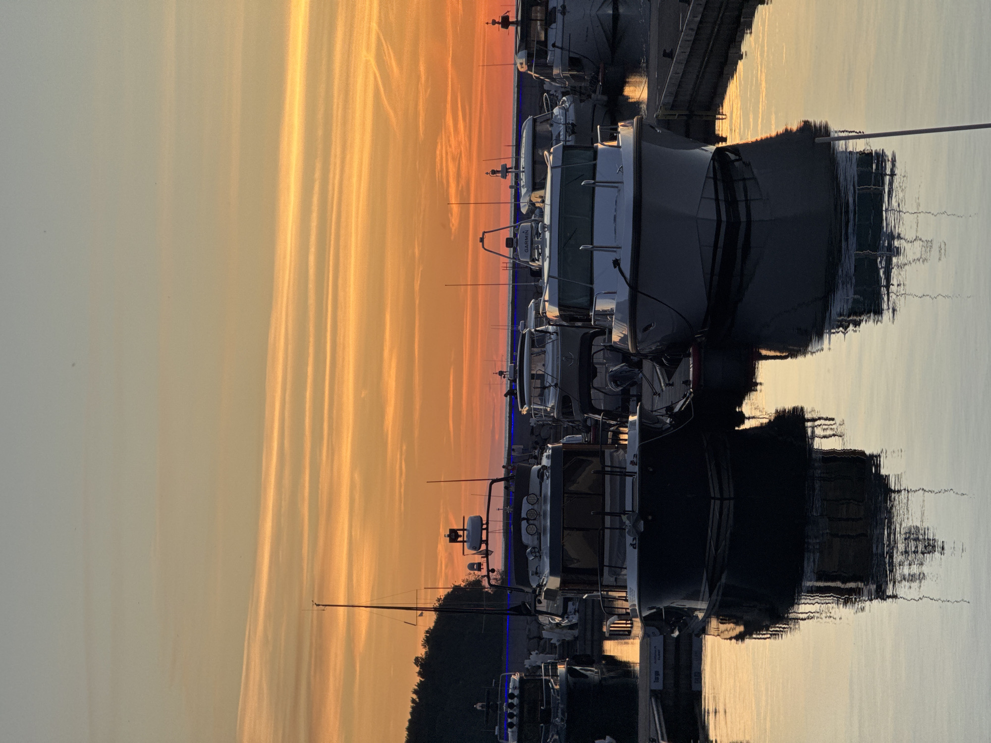 This image depicts a marina with several boats docked at sunset. The sky is filled with vibrant orange and yellow hues, indicating the sun is low on the horizon. The boats, mostly white and dark-hulled, are reflected in the calm water. One boat in the foreground has a "Garmin" logo visible on its radar. The scene is peaceful, with no people clearly visible in the immediate foreground or interacting with each other. The background shows a treeline and what appears to be a harbor wall. This scene is consistent with a location like the Tallinn Yacht Marina in Tallinn, Estonia, known for its scenic sunsets and numerous pleasure boats.