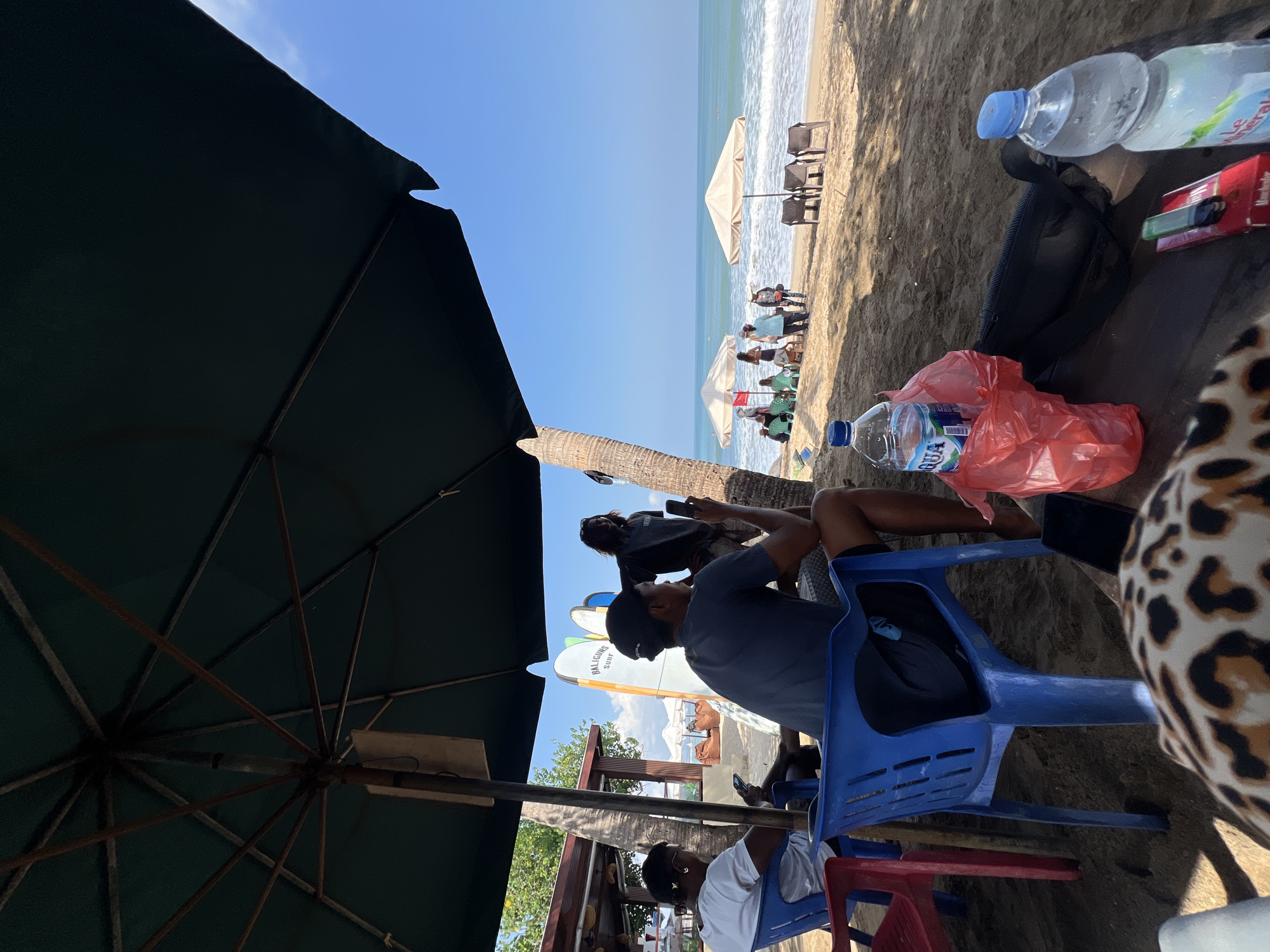 In Kuta, Indonesia, an outdoor scene unfolds on a sandy beach under a large, dark green umbrella. Two men are seated on blue plastic chairs, facing away from the camera. The man on the left wears a white t-shirt and sunglasses, holding a phone. The man on the right wears a dark t-shirt and a black baseball cap, also holding a phone. To the right, another man in a dark t-shirt and shorts stands near surfboards and a palm tree, looking at his phone. In the background, several other people are scattered across the beach, some sitting under beige umbrellas, while the ocean waves gently roll towards the shore. On a table in the foreground, a red plastic bag contains items, and two water bottles are visible, along with a pack of cigarettes and an e-cigarette. The sky is clear and blue.